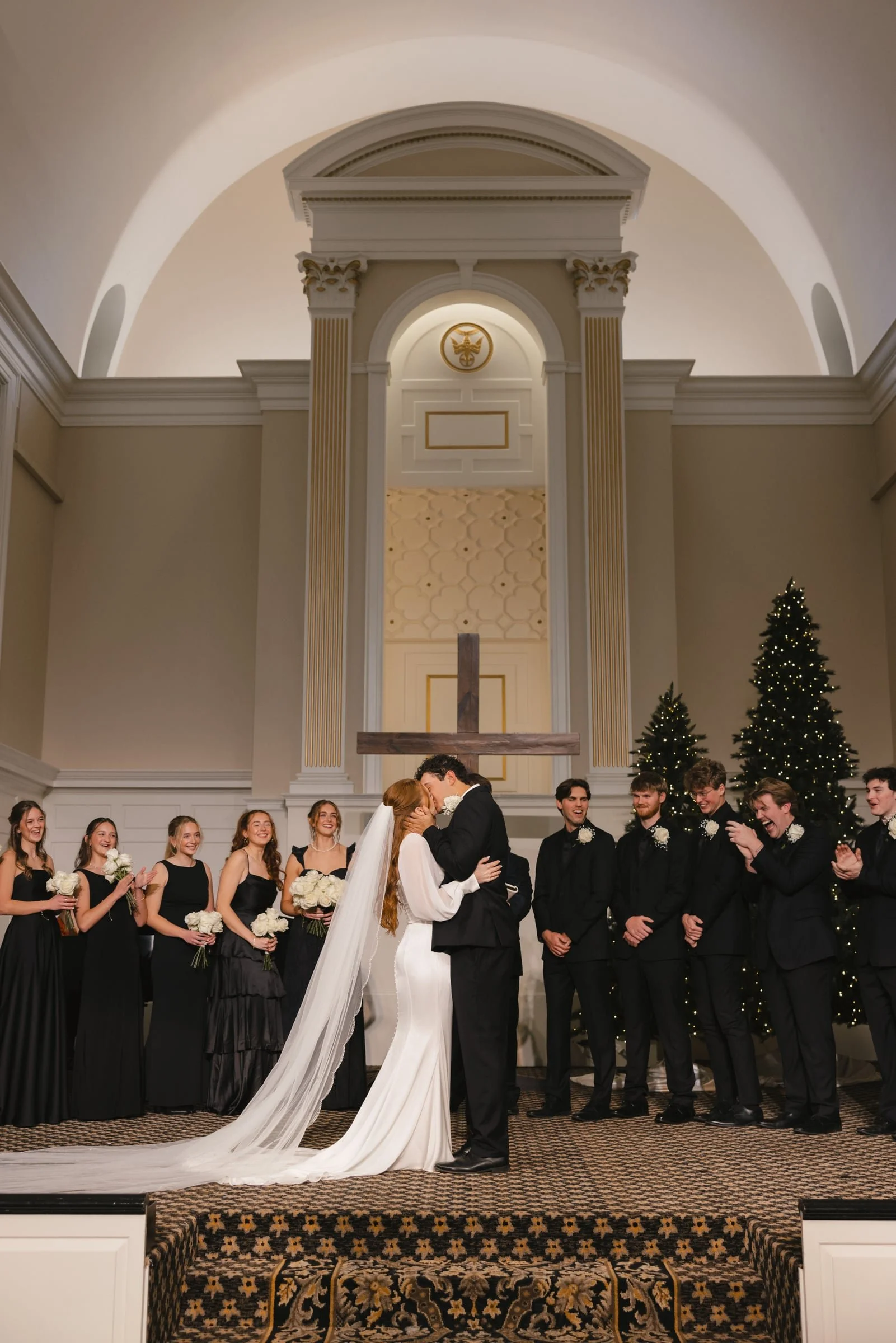 A wedding ceremony taking place in a church with a bride and groom kissing in front of a group of bridesmaids and groomsmen. The church has high ceilings, decorative columns, and Christmas trees with lights.