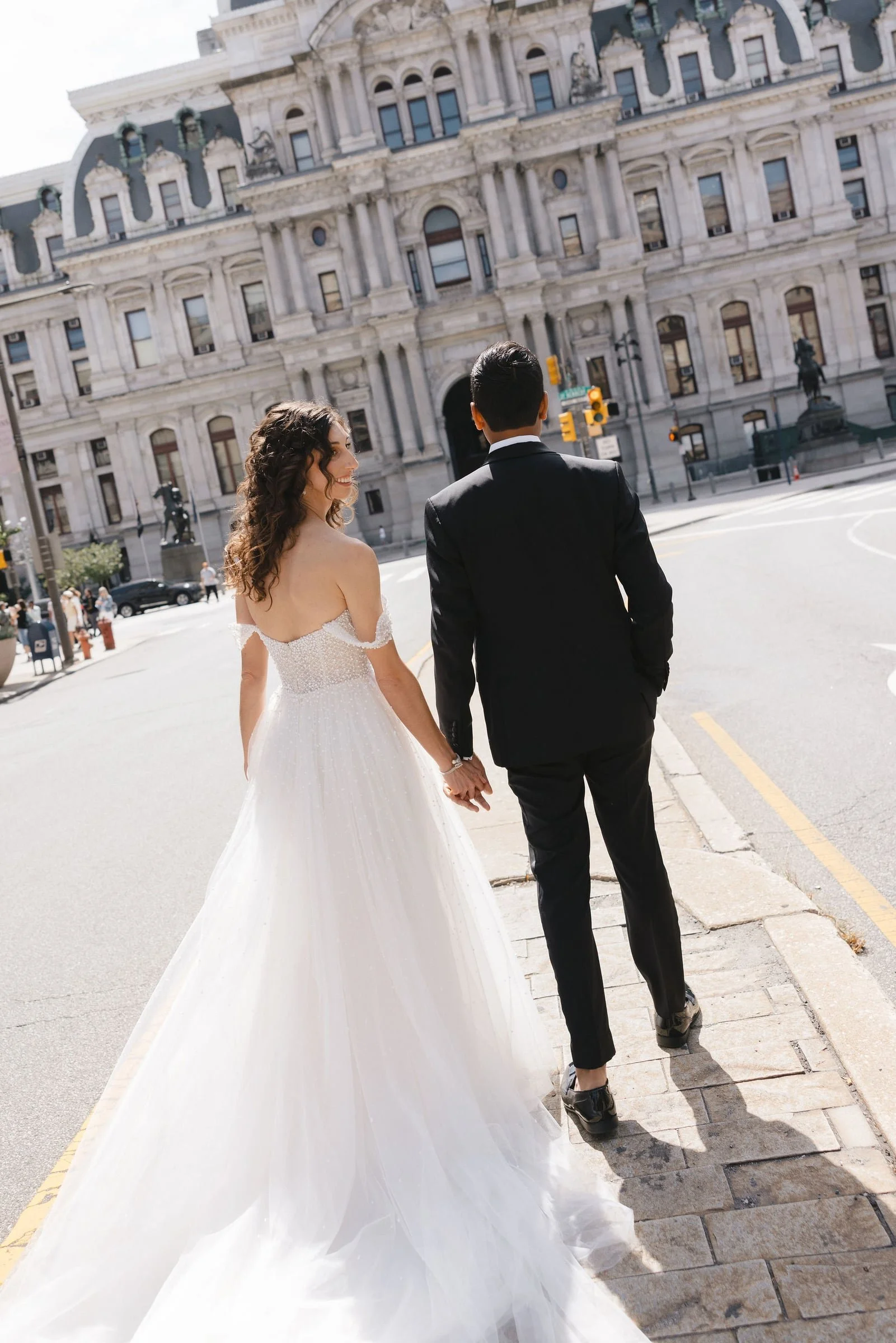 A bride and groom holding hands and walking in an urban area with historic buildings in the background. The bride is in a white wedding gown, and the groom is in a black suit. They are seen from behind.