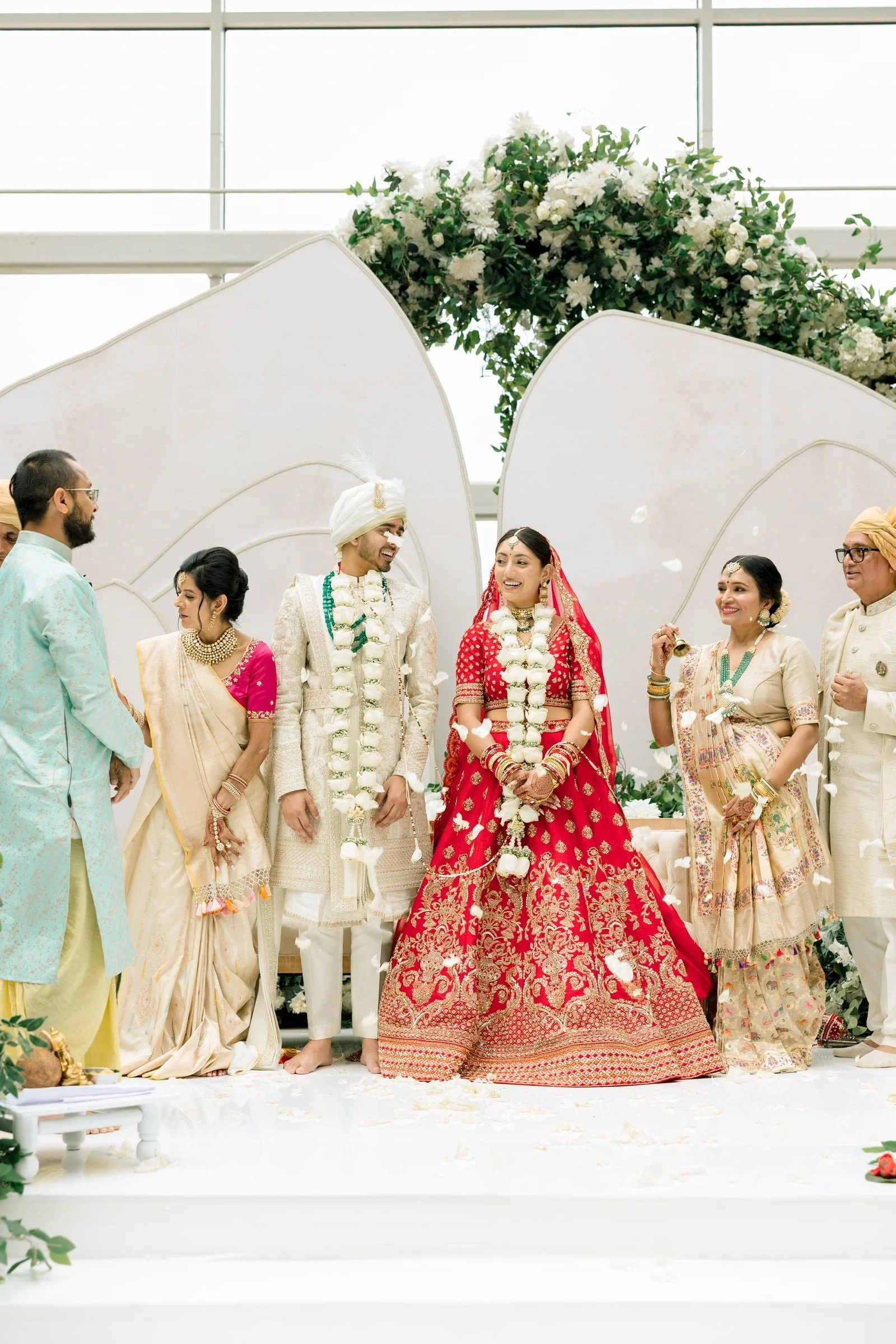 Indian wedding ceremony with the bride and groom standing together surrounded by family members, with white floral decorations and petals falling.