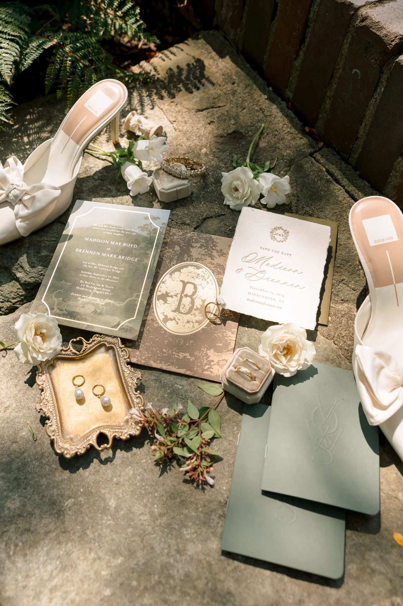 Wedding invitation, jewelry, flowers, and accessories displayed on a stone surface outdoors.