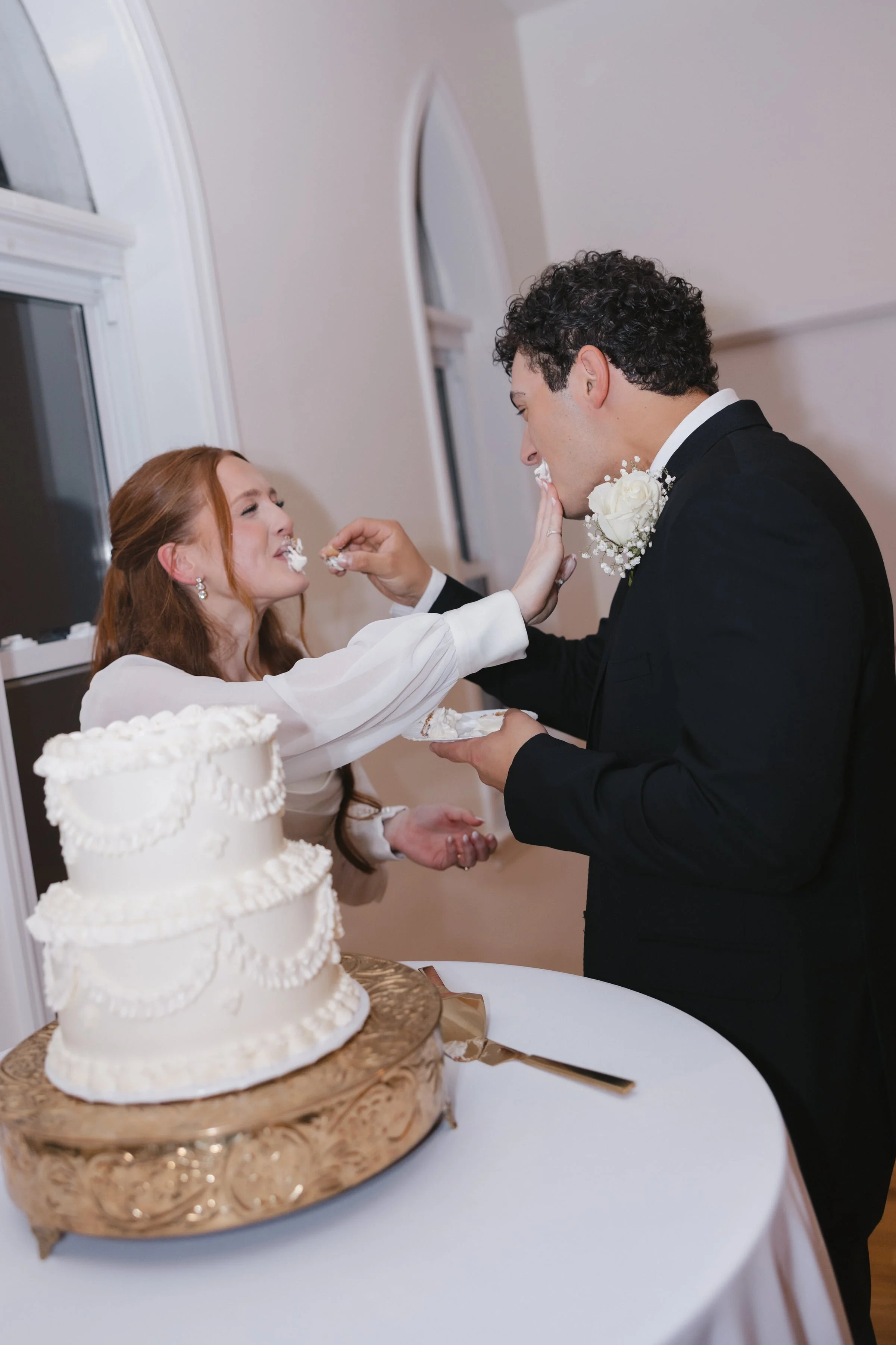 A bride and groom feed each other wedding cake at their wedding reception, with a three-tier white cake on a decorative stand in the foreground.