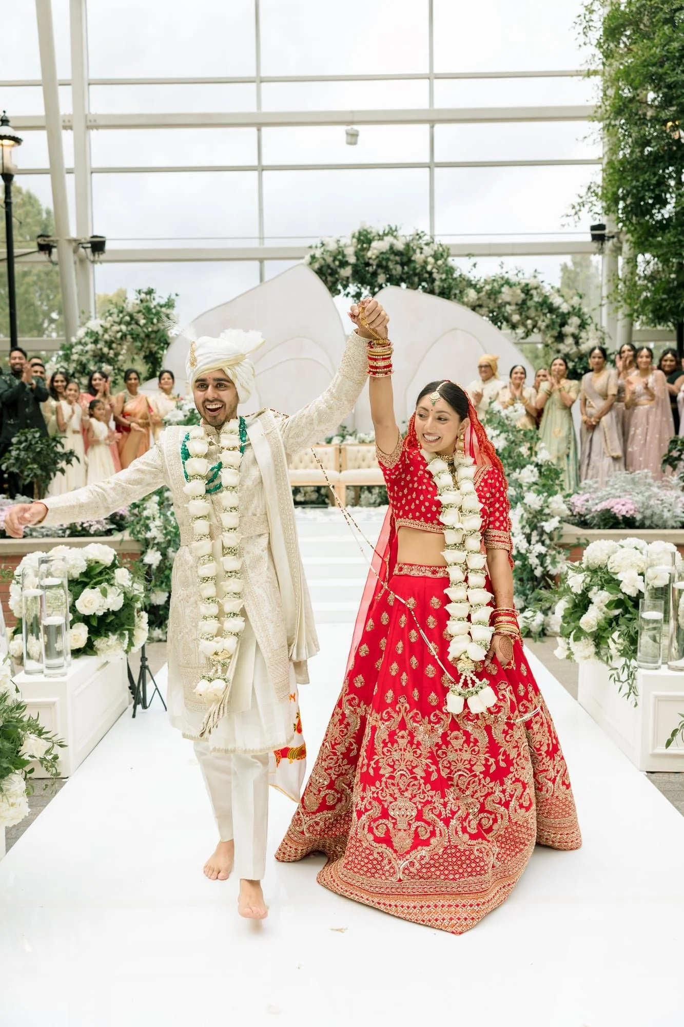 Indian bride and groom in traditional wedding attire celebrating and holding hands in a decorated venue with floral arrangements and guests in the background.