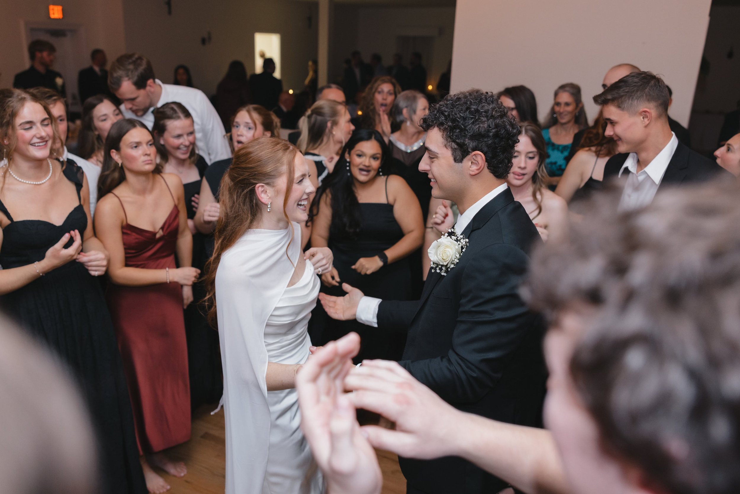 A bride and groom are dancing and smiling at their wedding reception, surrounded by guests who are also dancing and celebrating.