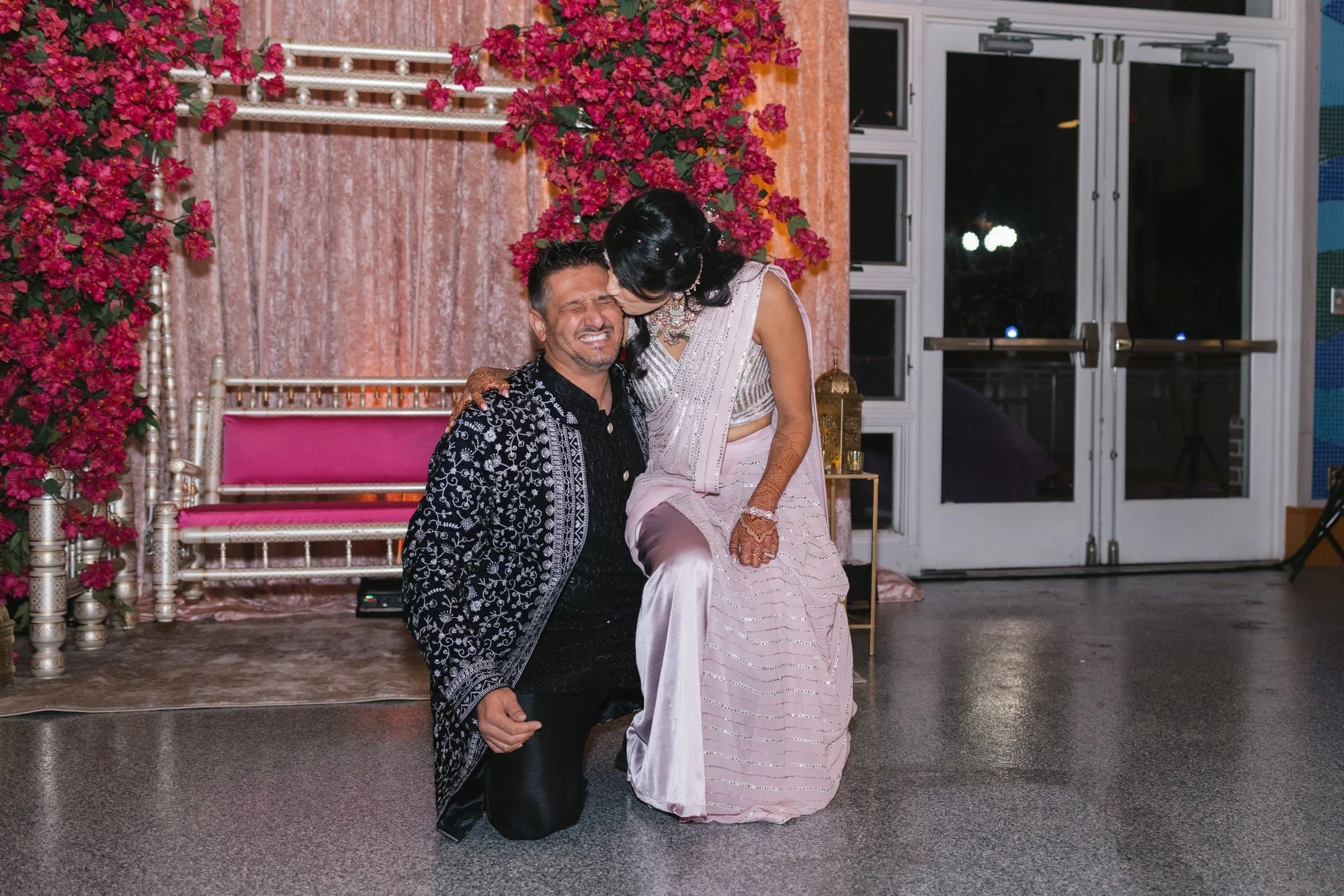 A man and a woman at a wedding reception. The man is kneeling and grimacing while the woman leans over and kisses his forehead. The woman is dressed in traditional Indian attire with henna on her hands. The background features pink floral decorations