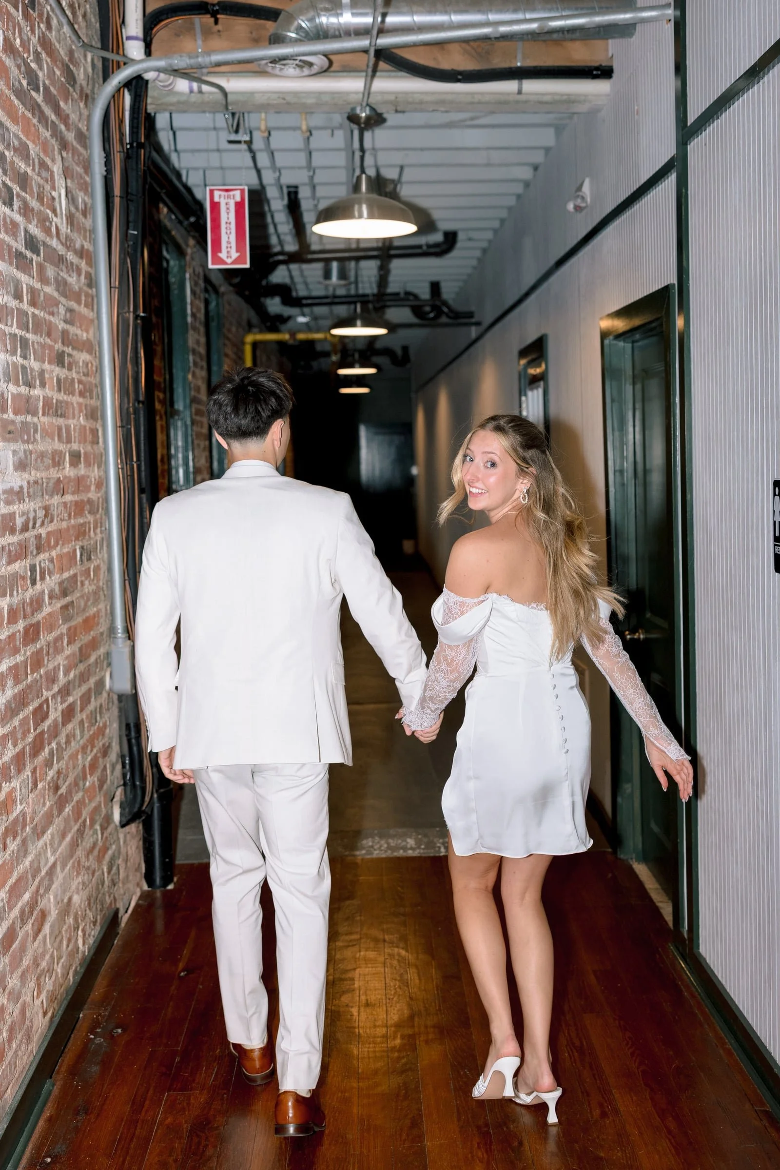 A man in a white suit holding hands with a woman in a white off-shoulder dress walking down an industrial-style hallway.