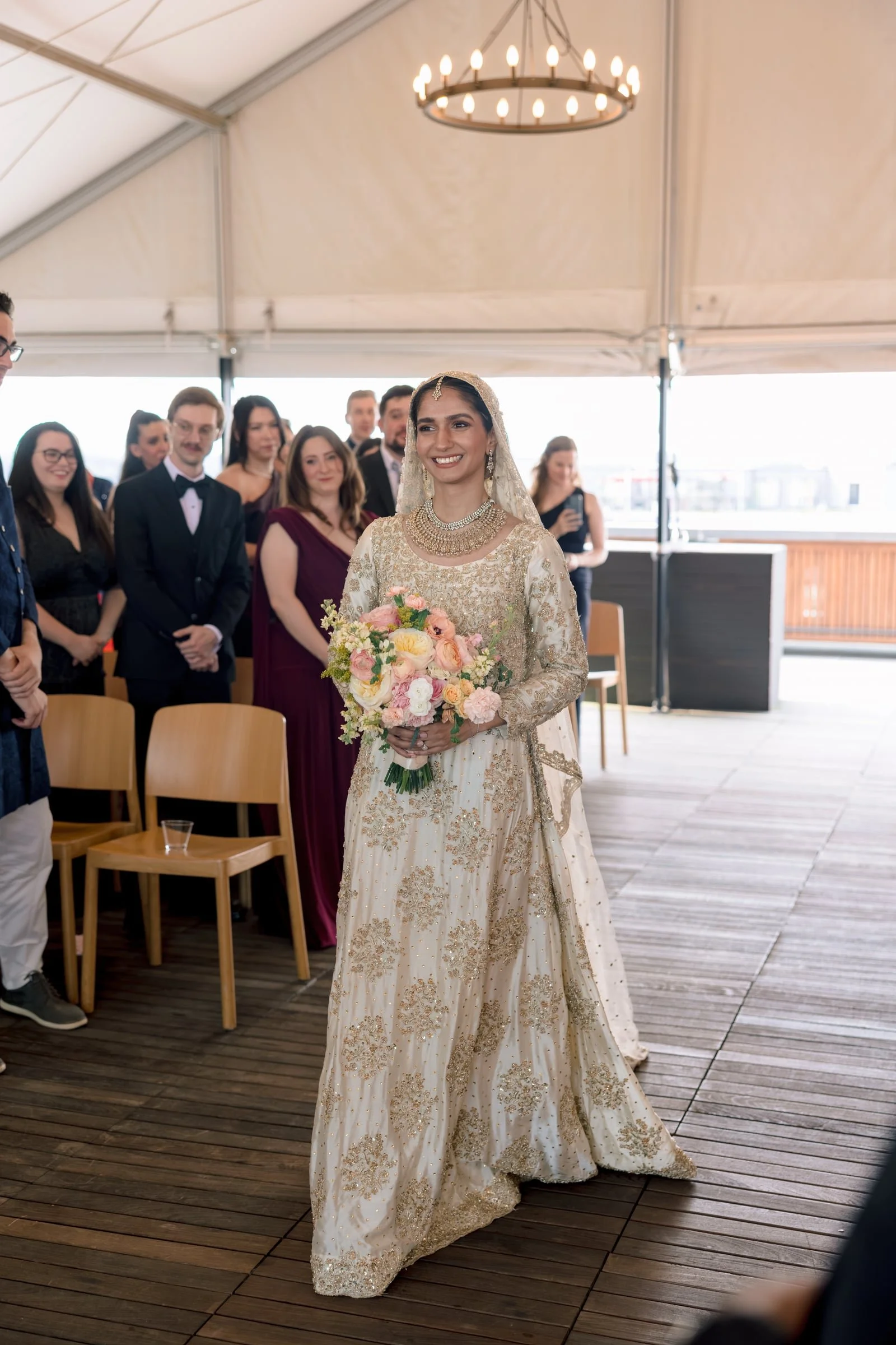 A bride in a traditional cream and gold embroidered wedding gown holding a bouquet of pink and white flowers during a wedding ceremony, surrounded by guests in formal attire inside a tent.