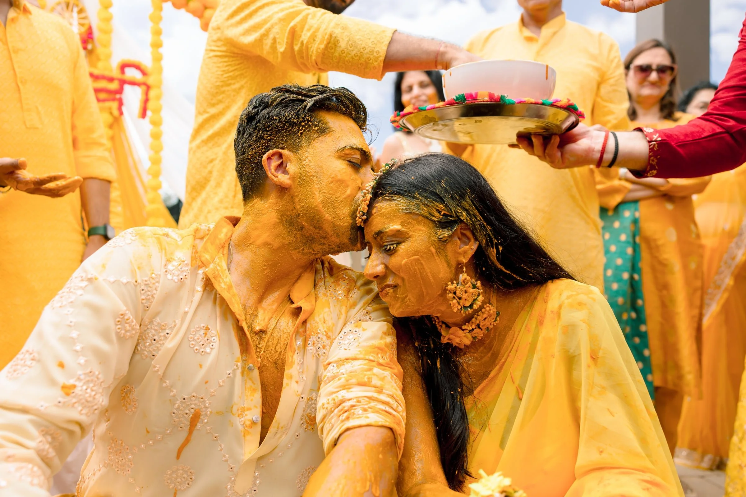 A couple participating in a traditional Indian celebration, with both covered in yellow powder, surrounded by people in yellow clothing, during a festive outdoor event.