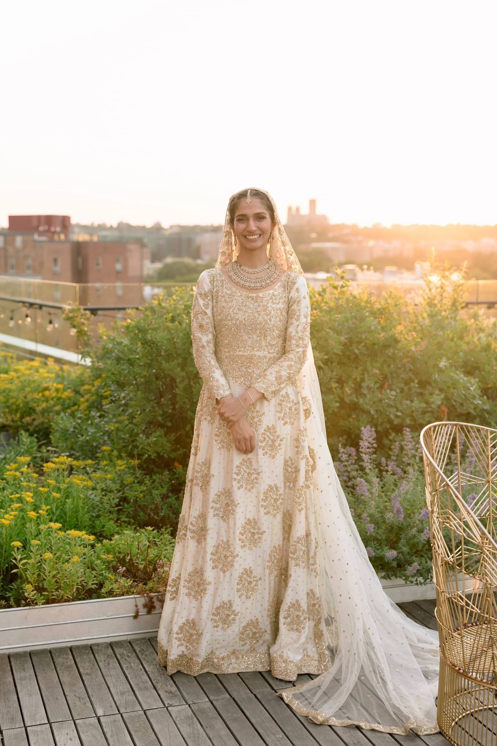 A woman wearing a traditional white and gold dress and accessories, standing on a wooden deck with a garden and city skyline in the background during sunset.