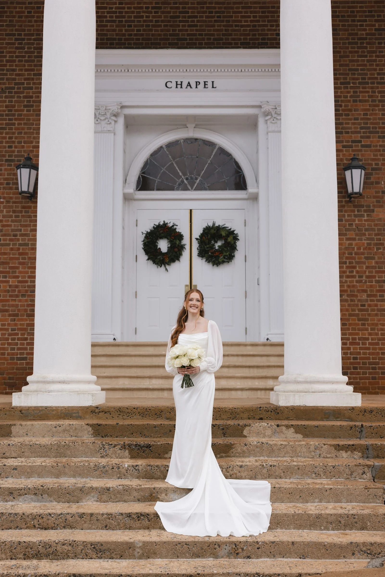 A smiling woman in a white wedding gown holding a bouquet of white roses, standing on the steps of a brick building with large white columns and a sign that says 'Chapel'.