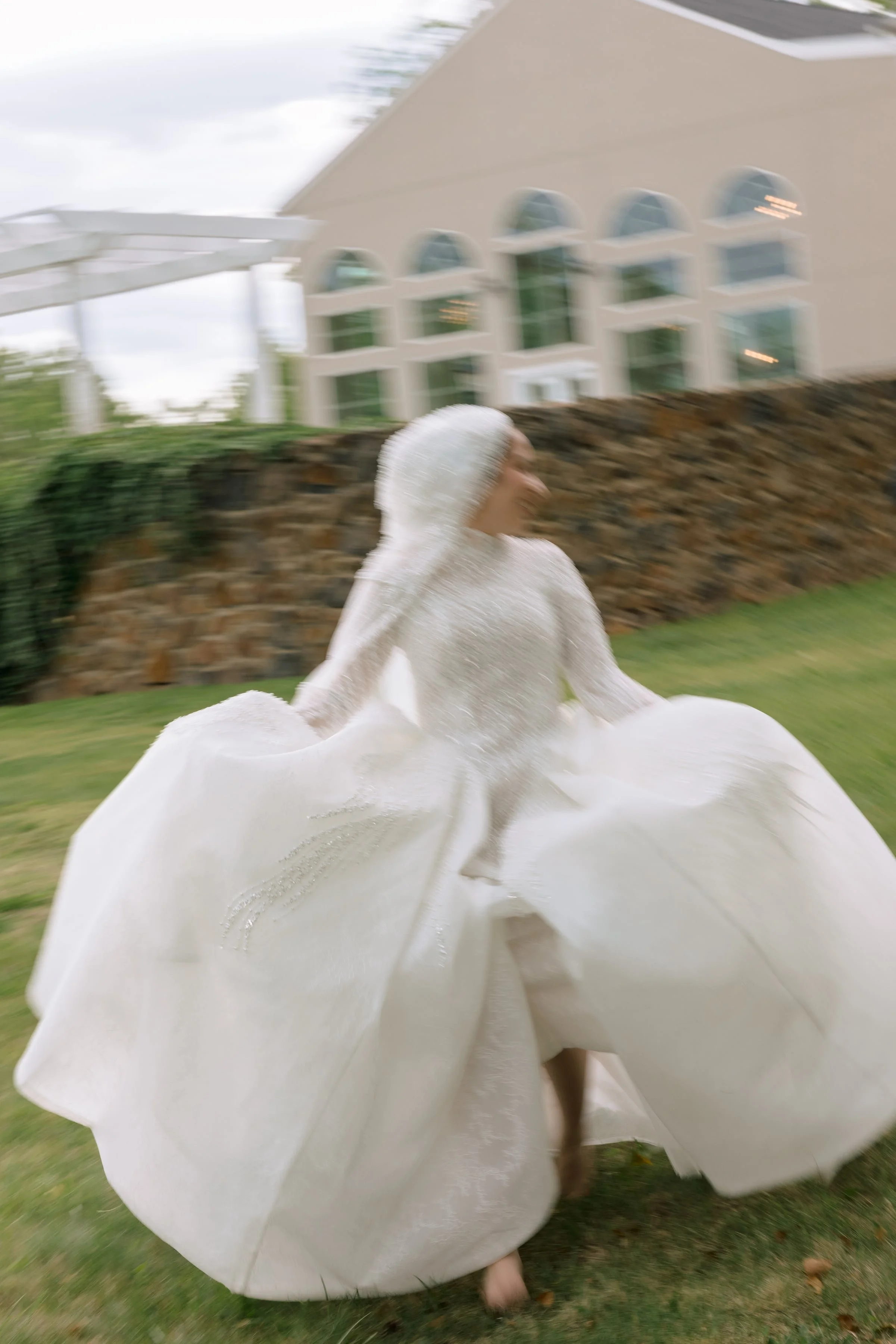 A person dressed in a white gown with a hood, resembling a wedding dress or a costume, is captured in motion outside near a stone wall and green grass. In the background, there is a beige building with large arched windows.