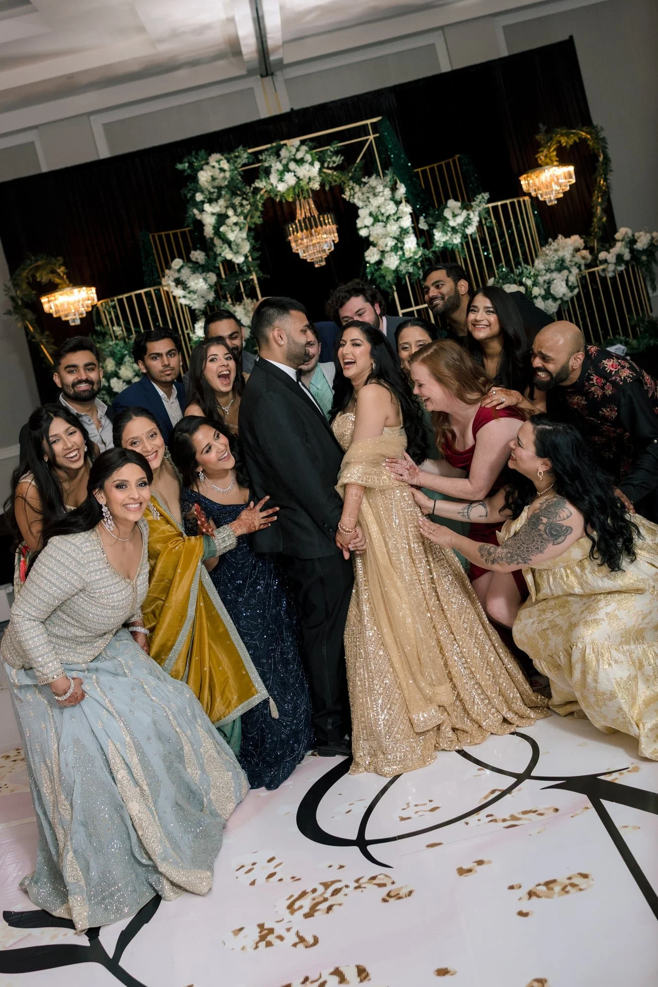 A group of people dressed in formal attire gathering at a celebration event, with a decorated floral backdrop and chandeliers.