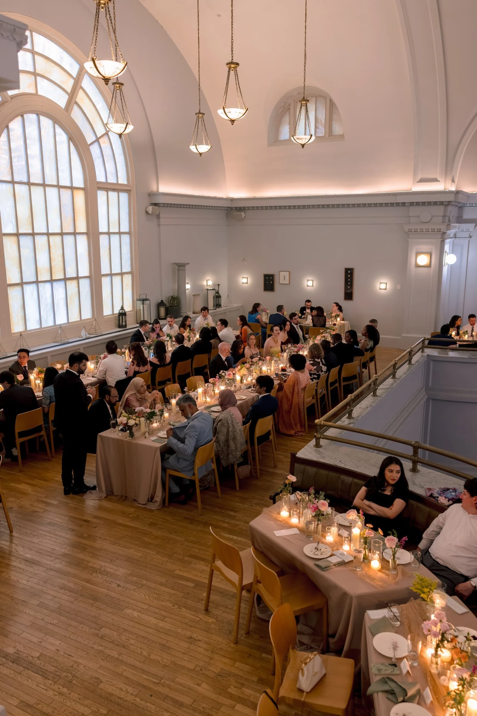 Indoor banquet hall with high arched windows letting in natural light, elegant ceiling lights, and long tables decorated with candles, flowers, and tableware during a formal event attended by many guests.