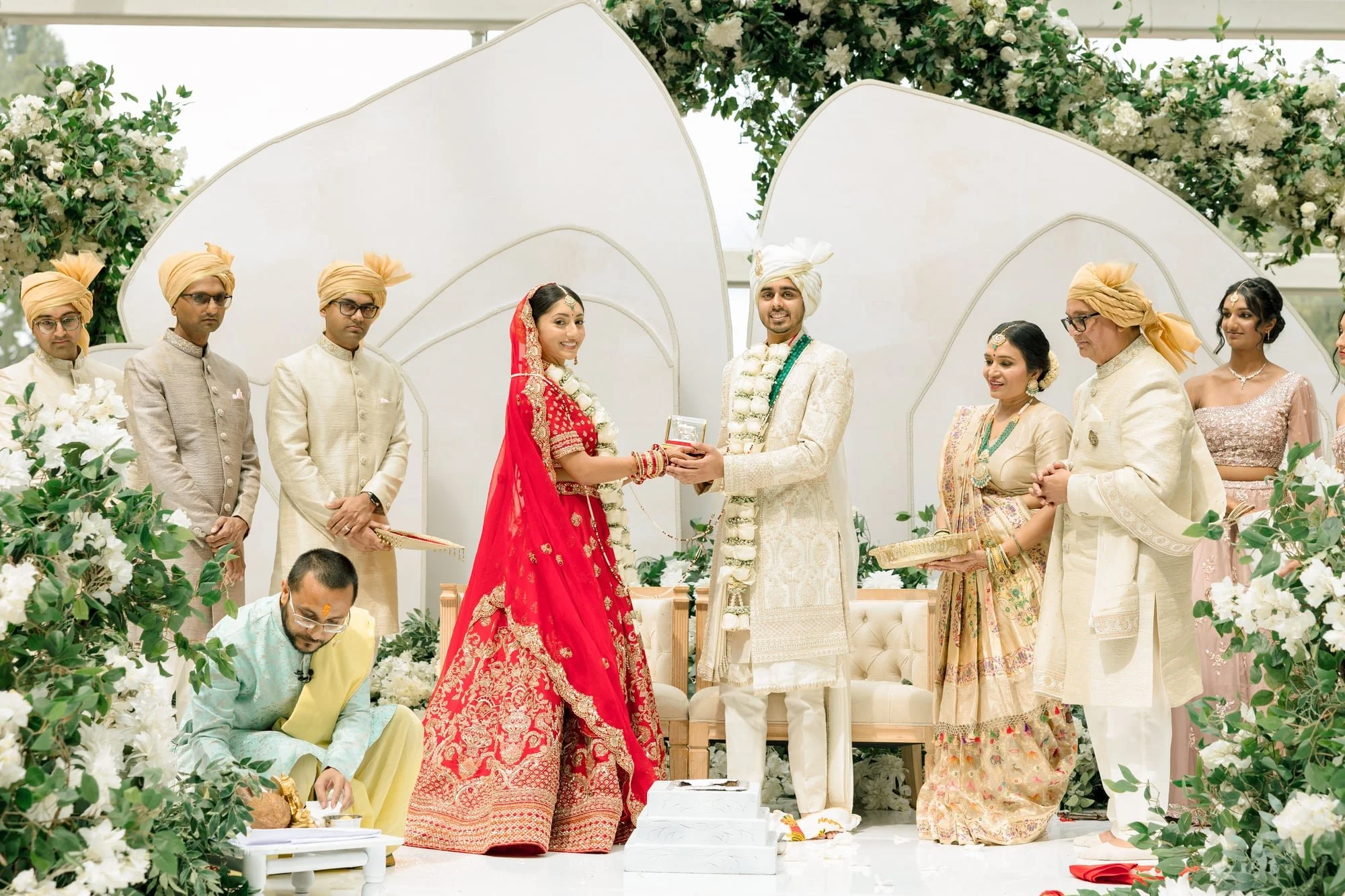 A traditional Indian wedding ceremony with the bride and groom standing on a decorated stage, surrounded by family members dressed in colorful attire. The bride wears a red and gold saree, while the groom wears a cream-colored sherwani and turban. Th