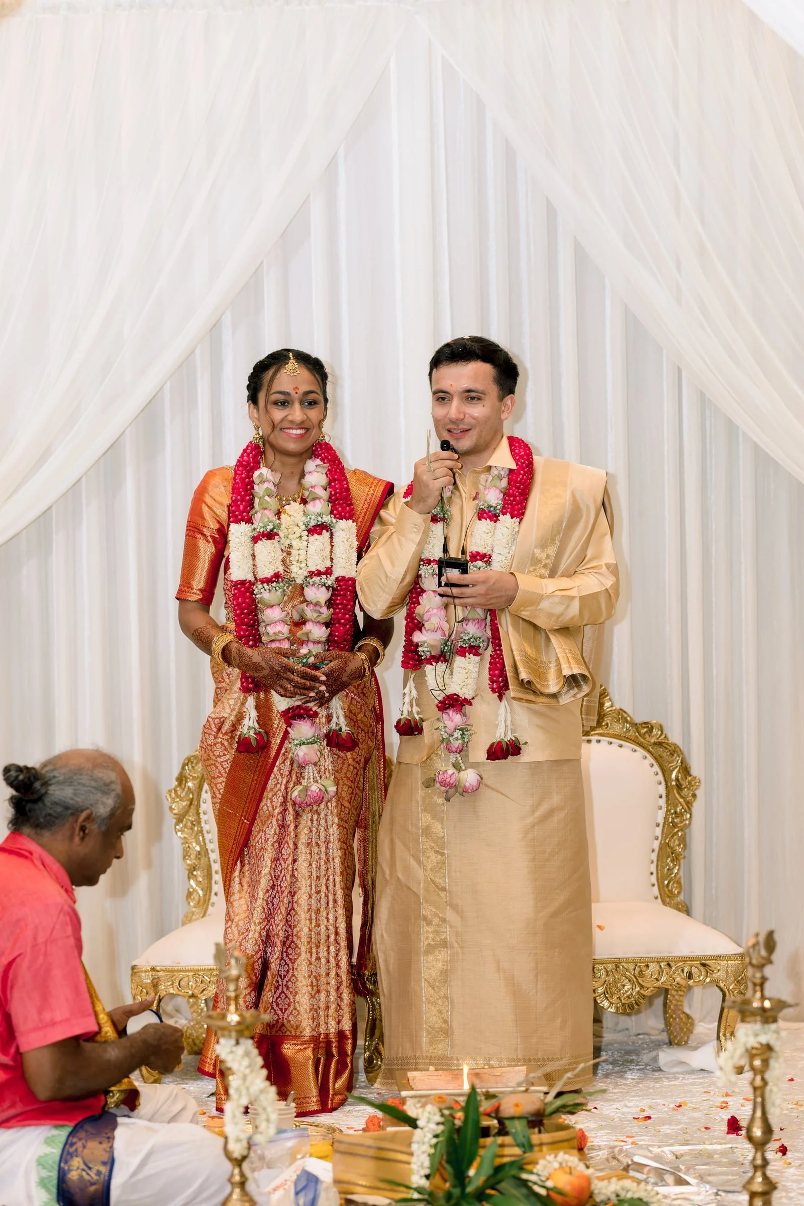 A traditional wedding ceremony with a couple standing on a decorated stage. The bride is wearing an orange saree and the groom is in a cream-colored traditional outfit. Both are adorned with floral garlands. There are people seated on the floor in fr