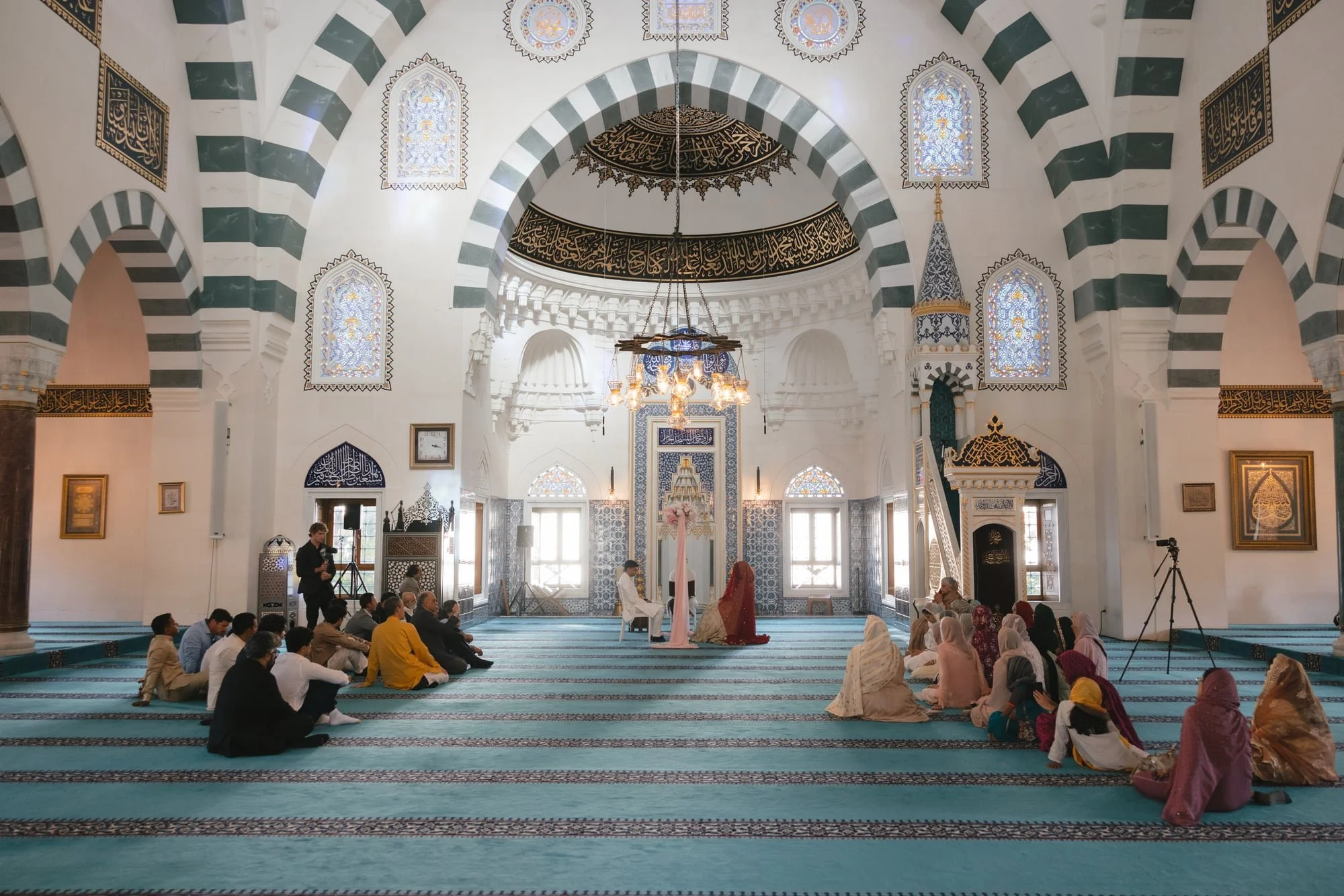 Interior of a mosque with people seated on a carpet, some on their knees, during a religious service or prayer. The mosque features arched windows with stained glass, intricate biblical calligraphy on the walls and ceiling, and a large chandelier han