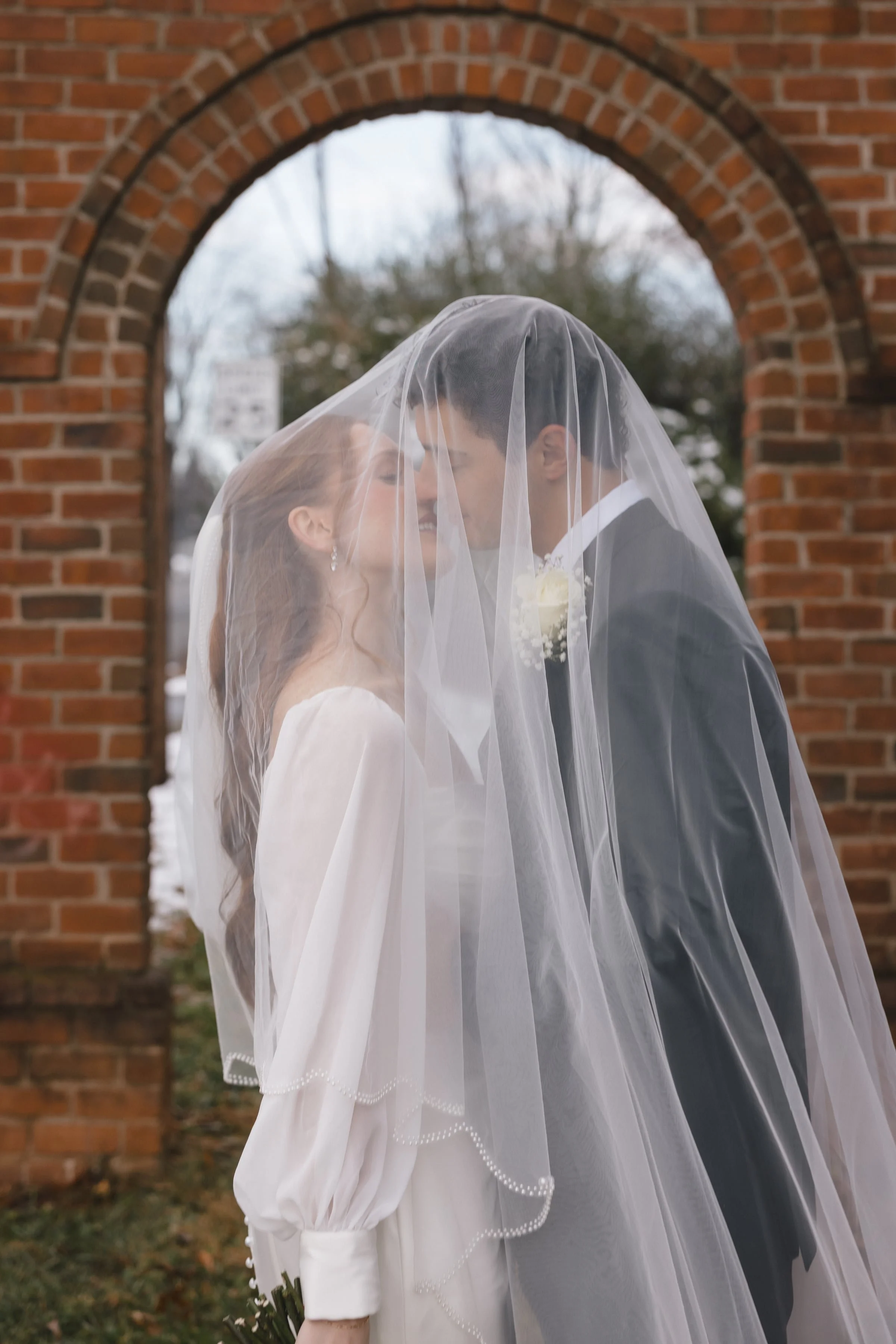 A bride and groom stand closely behind a sheer white veil, about to kiss, with a red brick archway in the background on an outdoor setting. The bride has light brown hair and is wearing earrings, while the groom has dark hair and is dressed in a tuxe