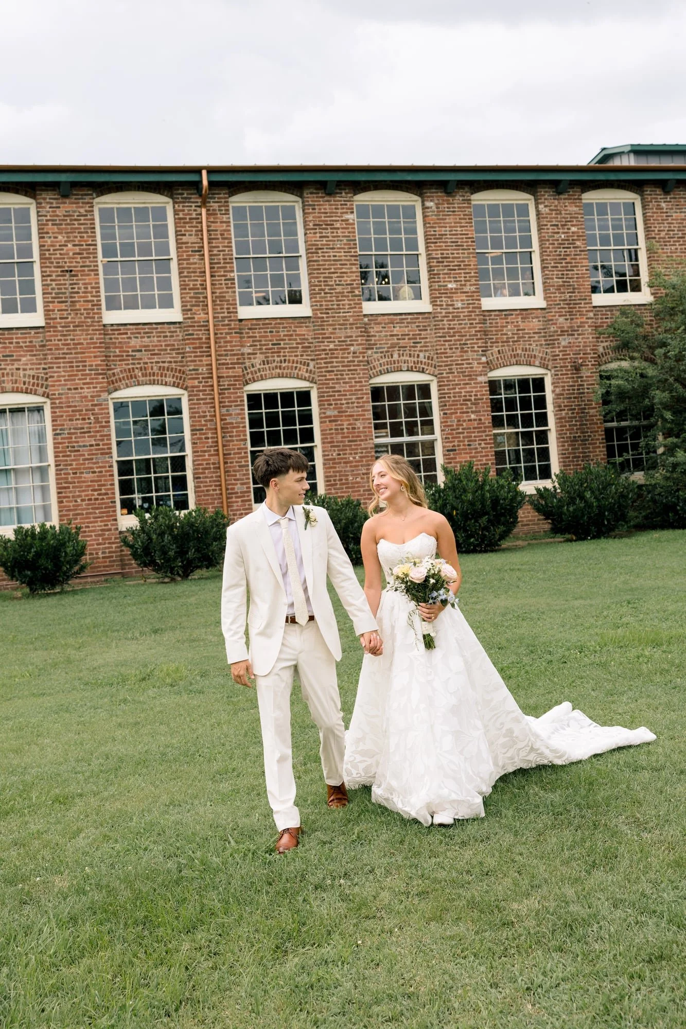 A bride and groom walking hand in hand on a lush green lawn in front of a large brick building with multiple windows, during a wedding celebration day.