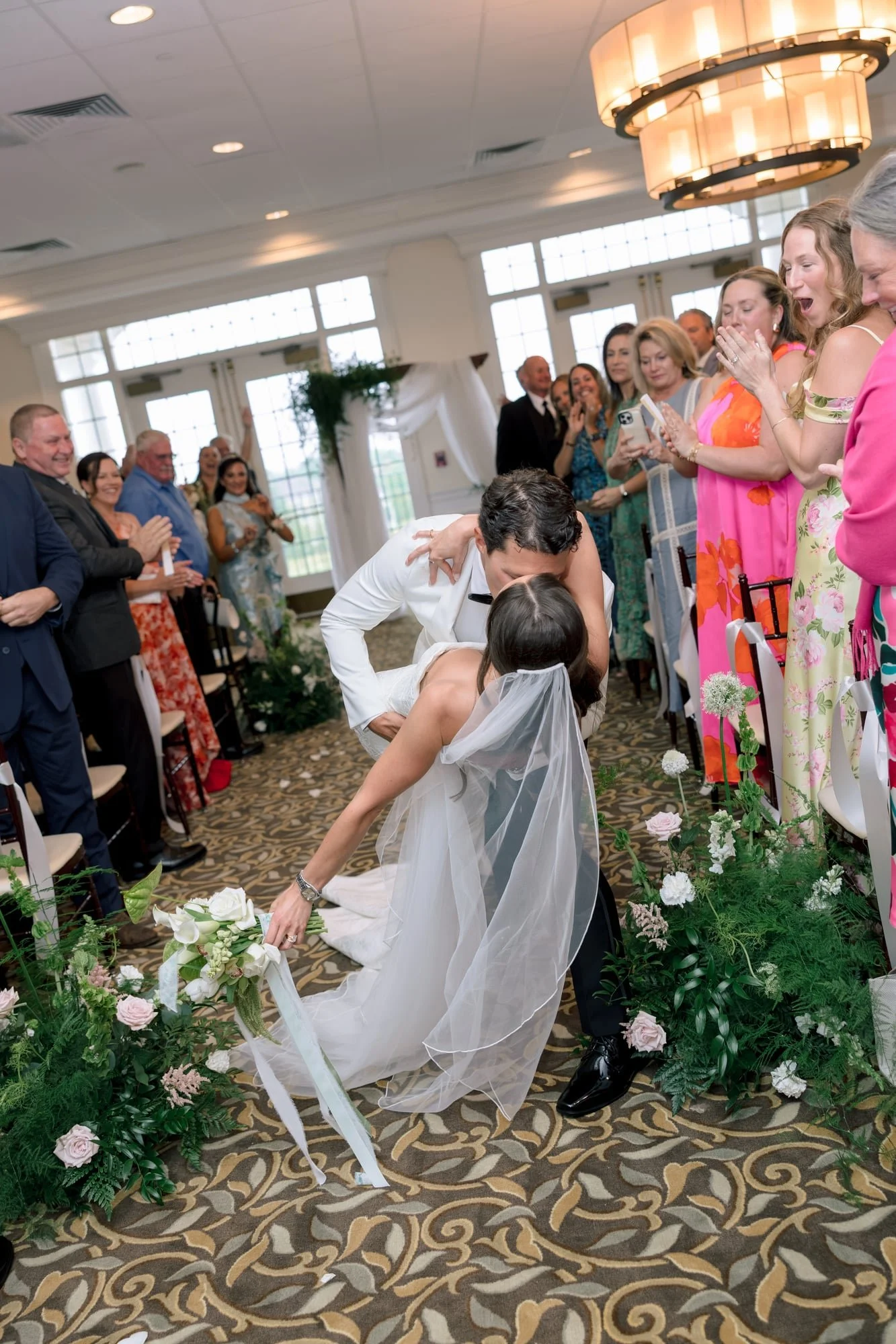 Bride in white dress and veil kneeling down to pick up her bouquet while groom leans over her during a wedding ceremony, with guests watching and smiling, in a decorated indoor reception hall with large windows and floral arrangements.