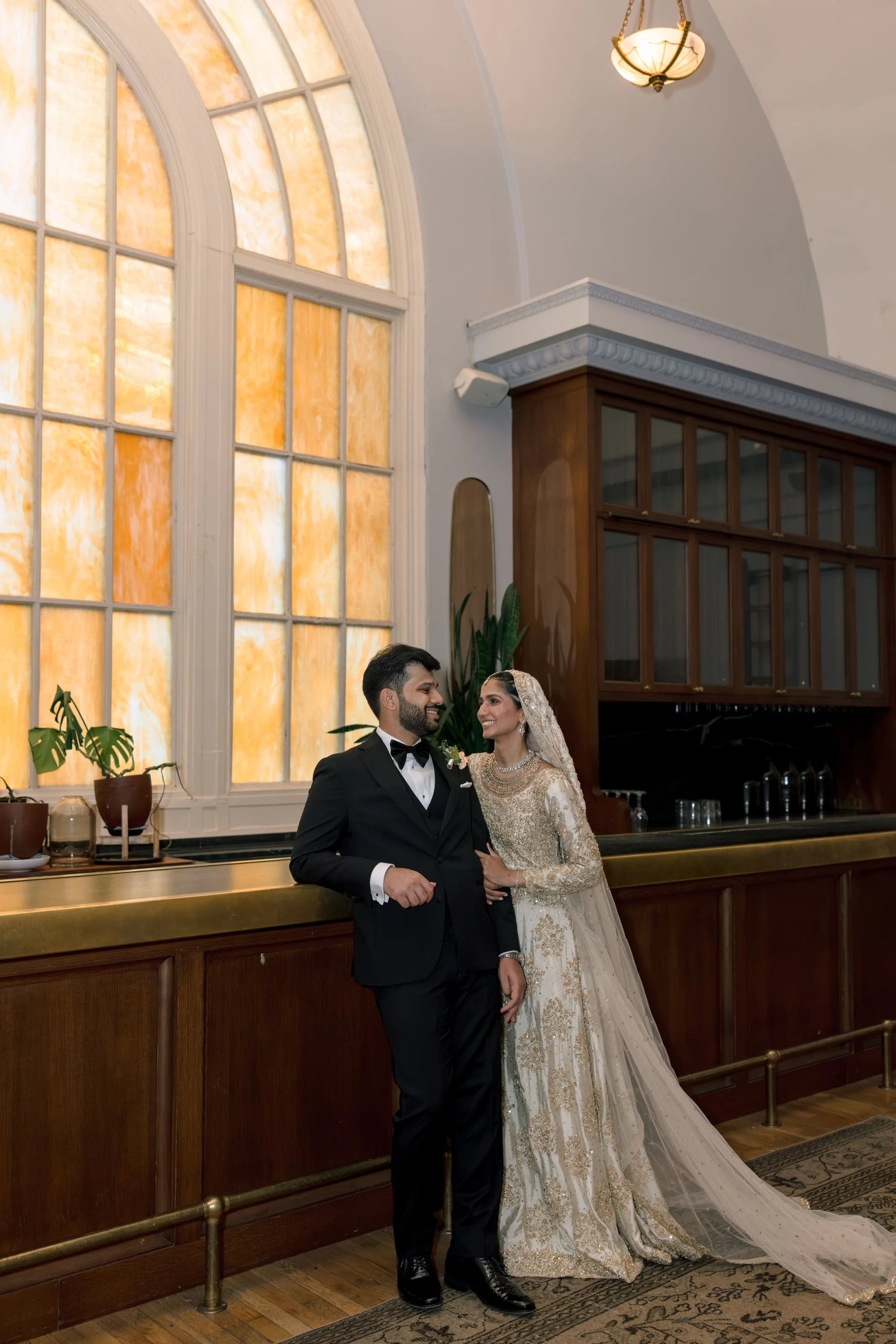 A newlywed couple dressed in wedding attire standing inside a grand room with tall stained glass windows, a wooden bar, and elegant lighting, smiling at each other.