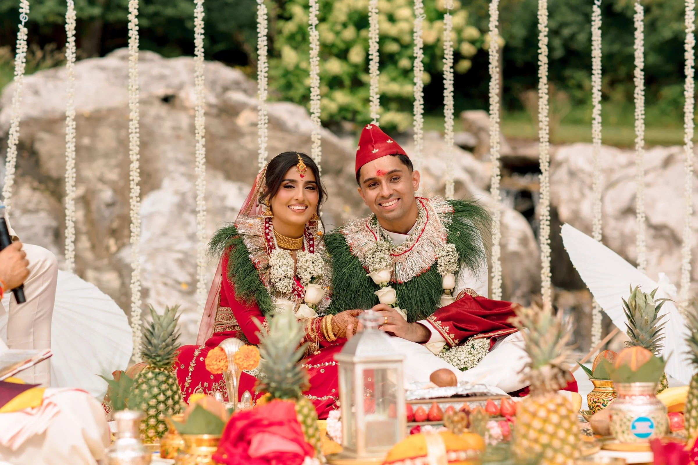 A newlywed couple dressed in traditional Nepali wedding attire sitting together outdoors during their wedding ceremony. The woman wears red and gold jewelry with a red sari, and the man wears a red topi and traditional clothing. Decorations include hanging white flowers and various fruits on the table in front of them.