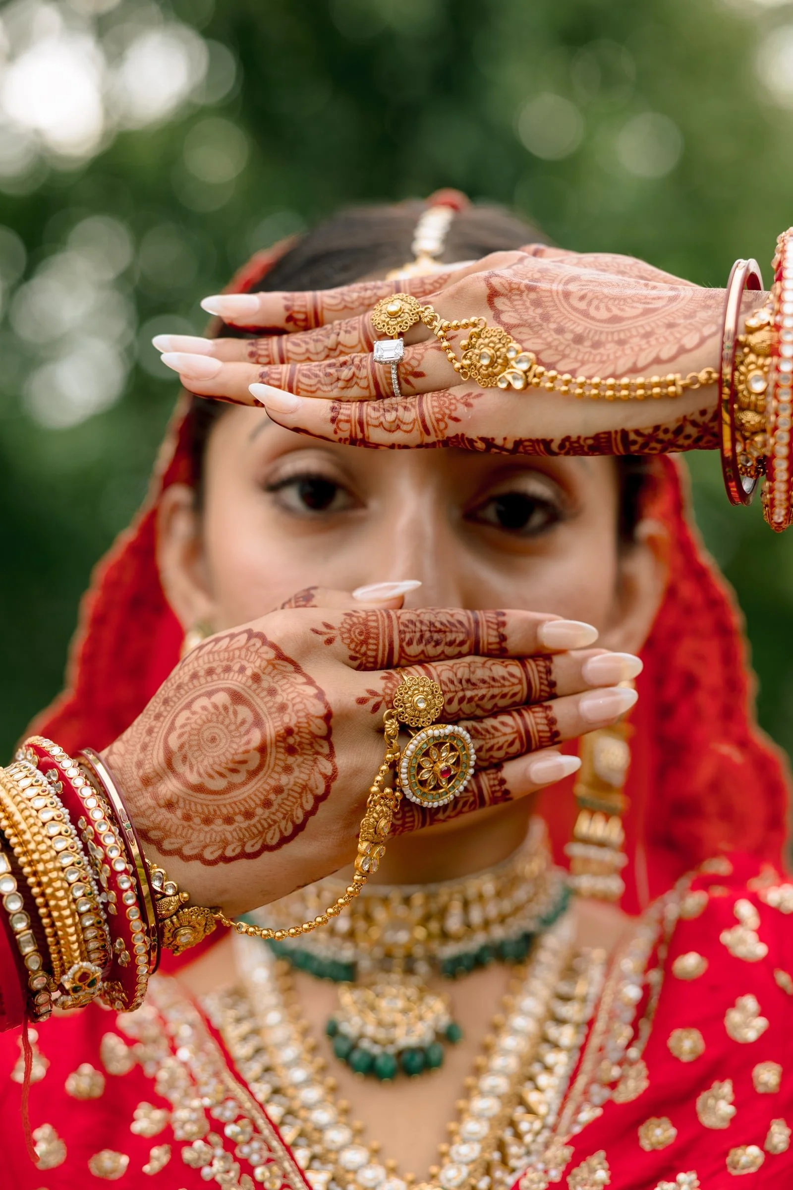 Close-up of a woman in traditional Indian bridal attire showing detailed red mehndi hand art, jewelry including rings, bangles, necklaces and earrings, with a blurred green outdoor background.