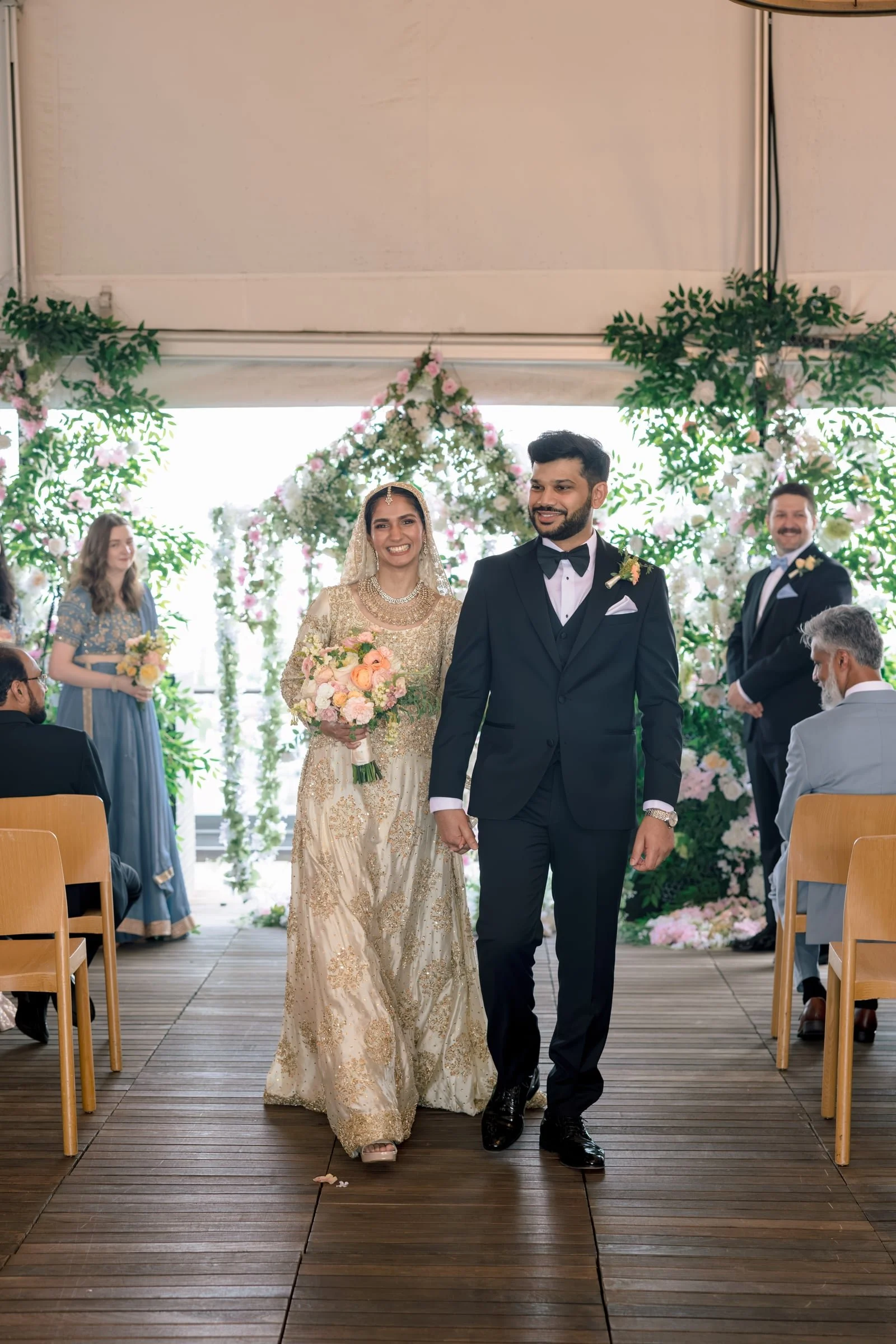 A bride and groom walking down the aisle at a wedding ceremony decorated with flowers and greenery, with guests seated on either side.