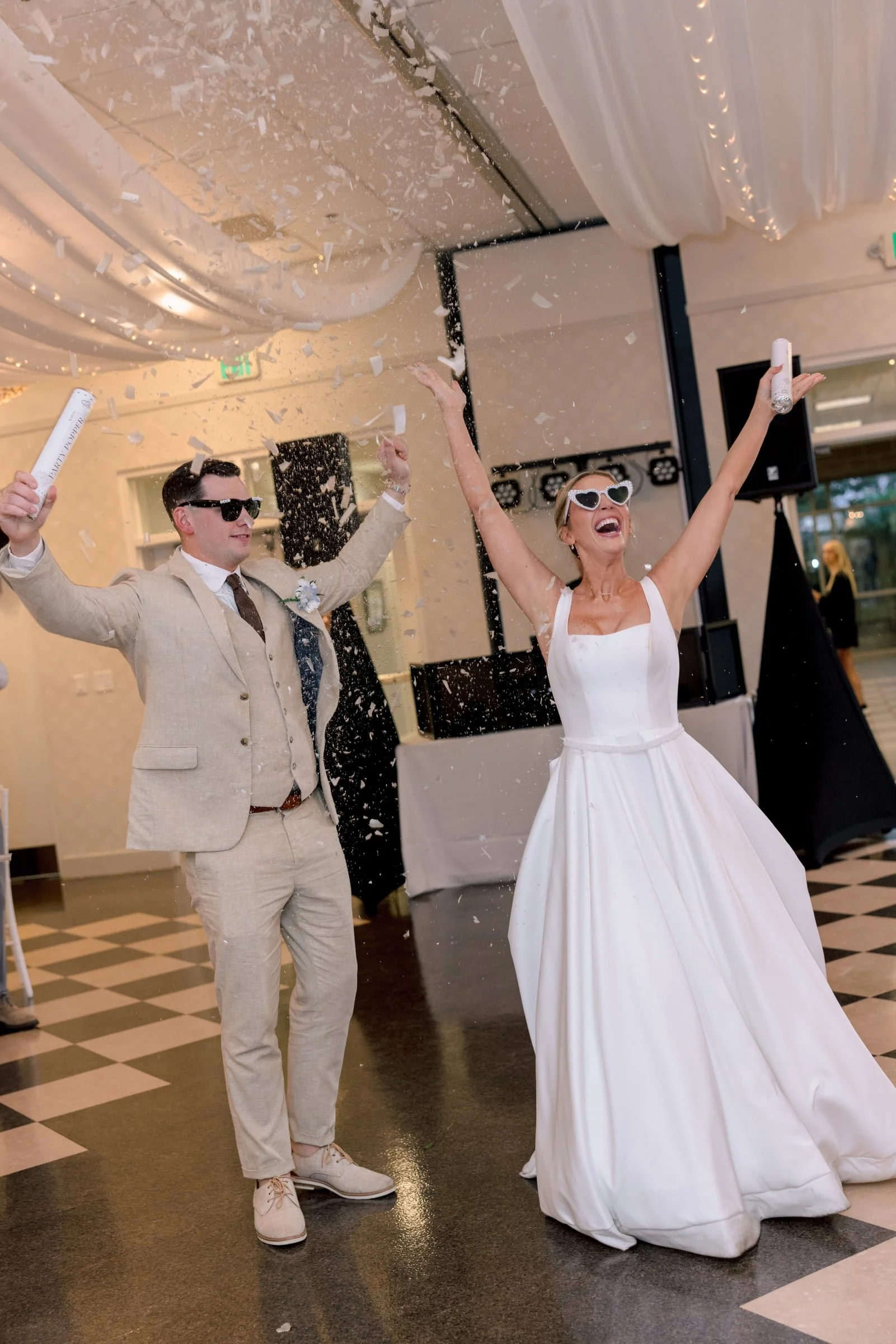A bride and groom celebrating with confetti at their wedding reception indoors. The bride is wearing a white wedding dress and white sunglasses, raising her arm happily. The groom is dressed in a beige suit, white shirt, brown tie, and sunglasses, al