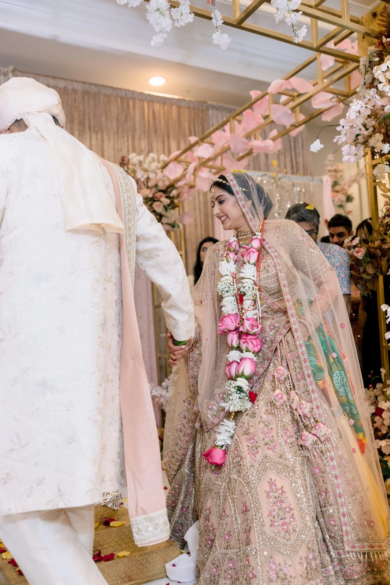 A bride in a traditional colorful bridal dress and a groom dressed in white traditional attire at a decorated wedding venue with floral arrangements and gold accents.