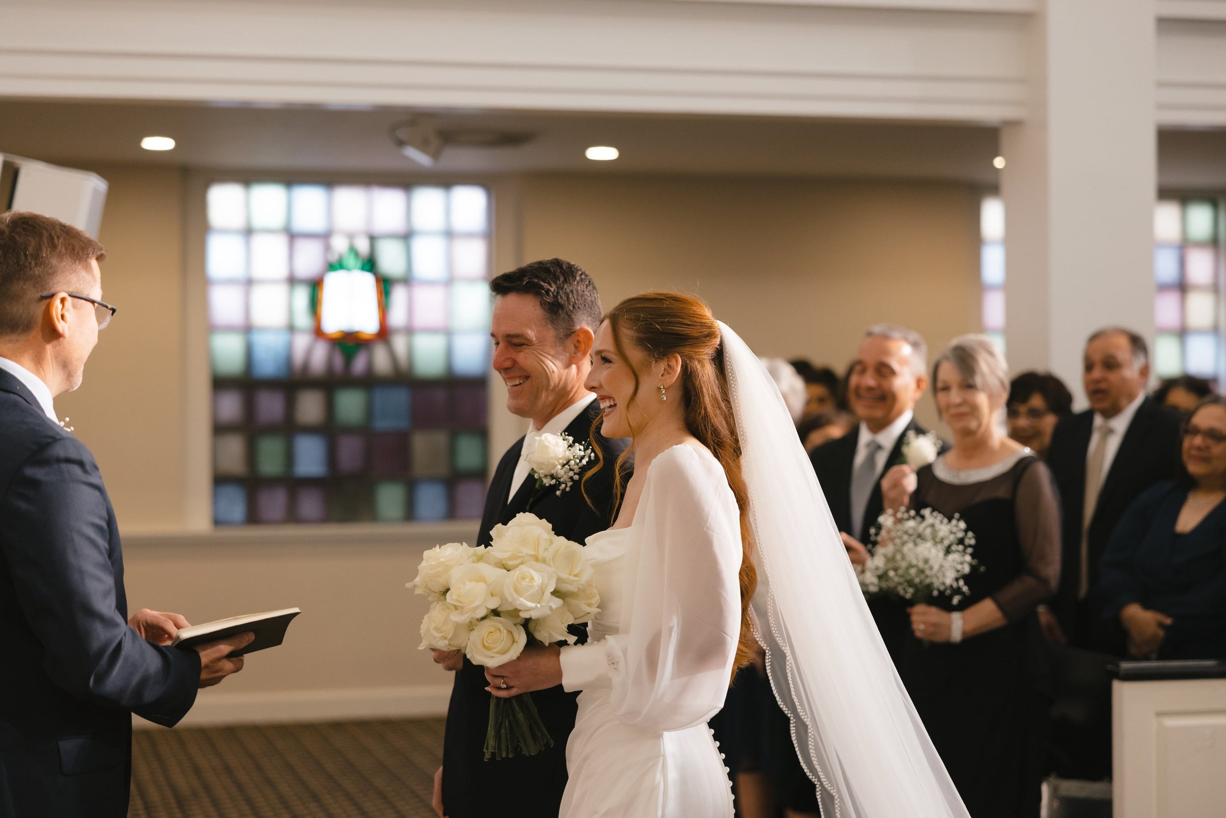 A bride and groom exchanging vows in a church during their wedding ceremony, with guests attending and a stained glass window in the background.