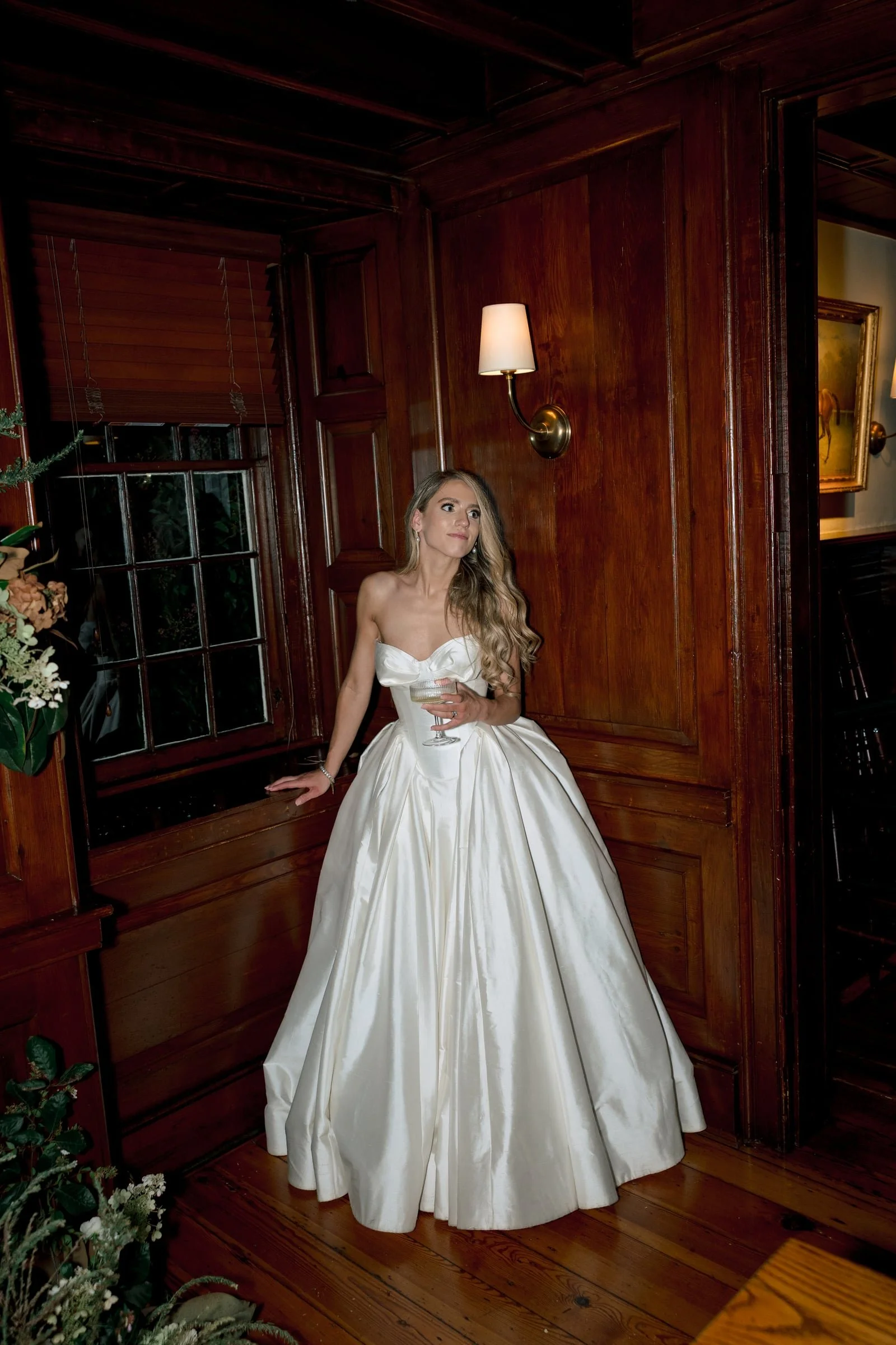 A woman in a strapless white gown with a full skirt is holding a glass of champagne and leaning against a wooden wall in a warmly lit room. The room has wooden paneling, a window with blinds, and wall-mounted light fixtures.