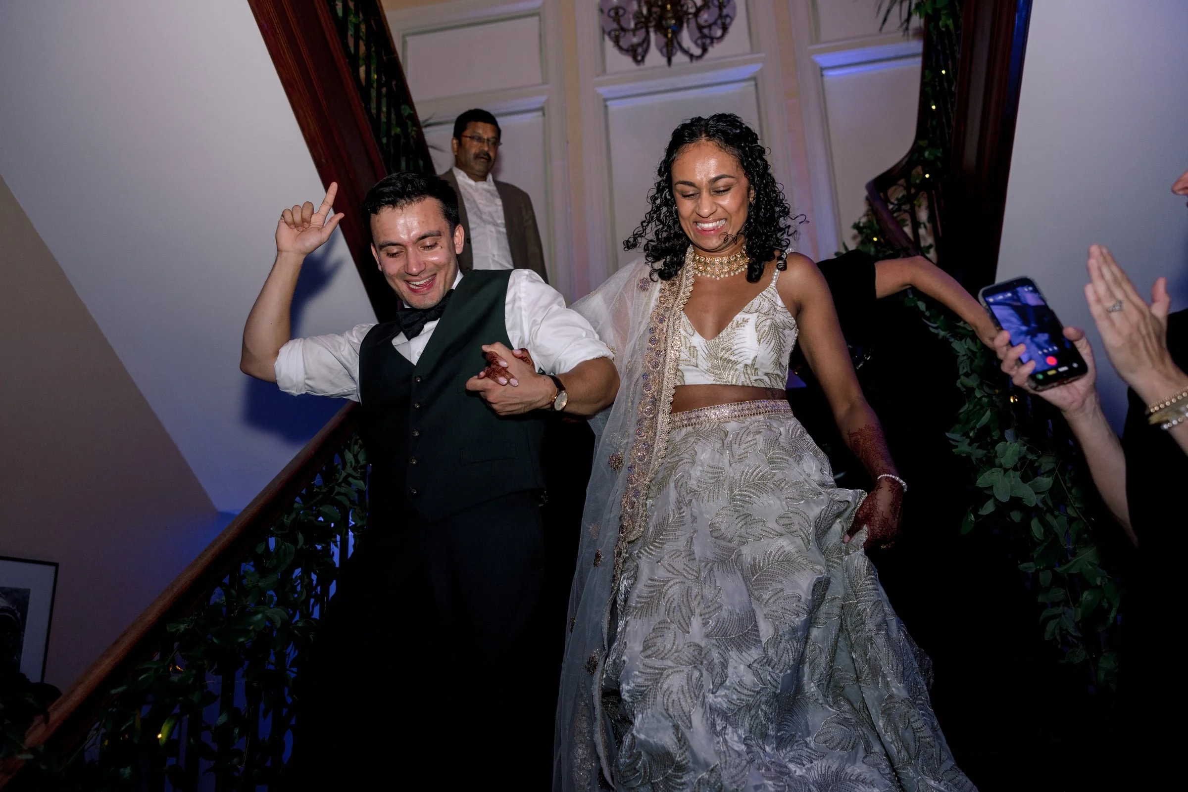 A joyous wedding reception scene showing a bride and groom dancing on a staircase, with a man in a suit watching, and a person holding a smartphone capturing the moment.