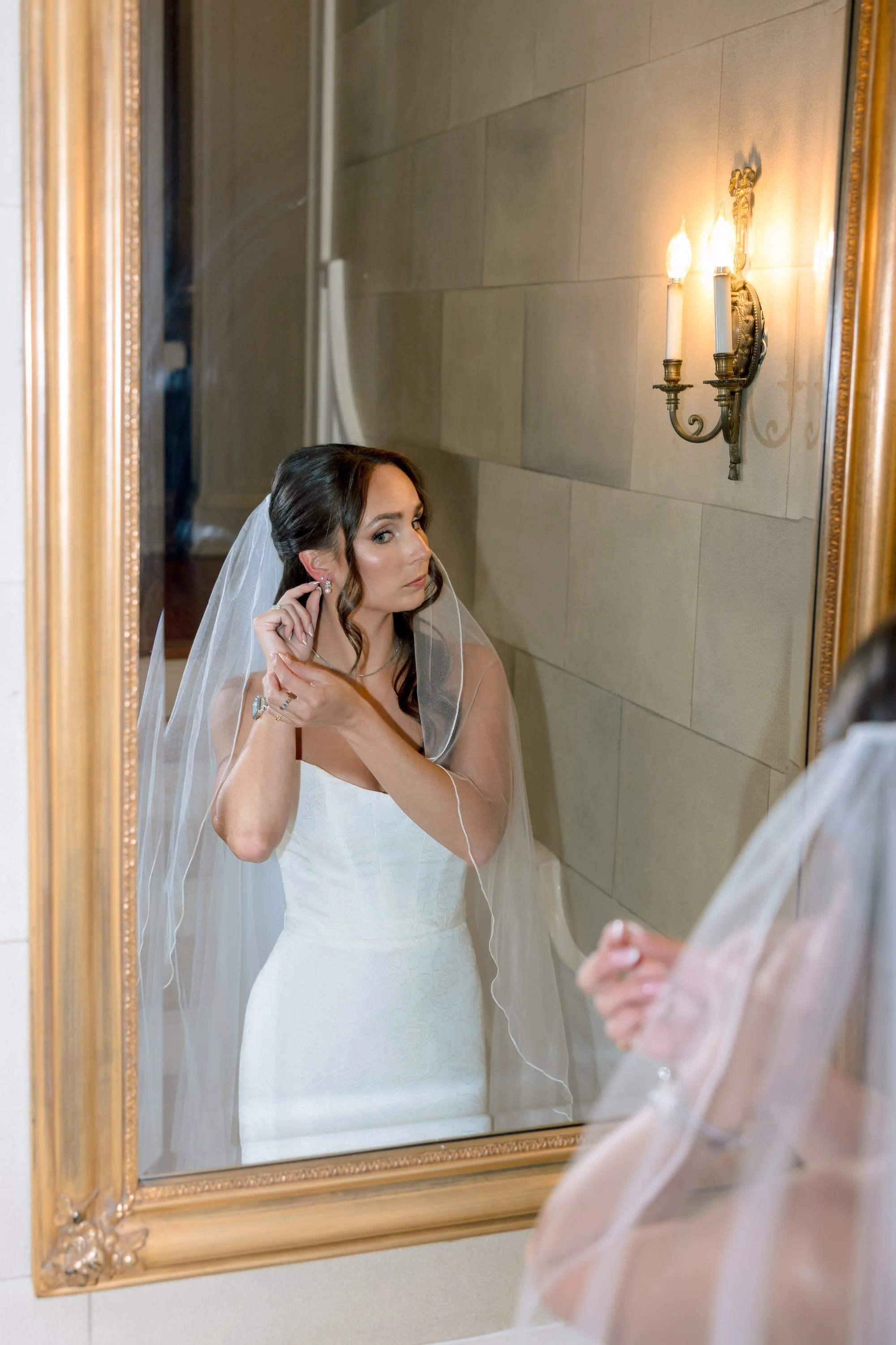 A bride with wavy brown hair and a veil is putting on earrings while looking in a large ornate gold mirror in a room with beige tiled walls and a wall sconce with two candle-style lights.