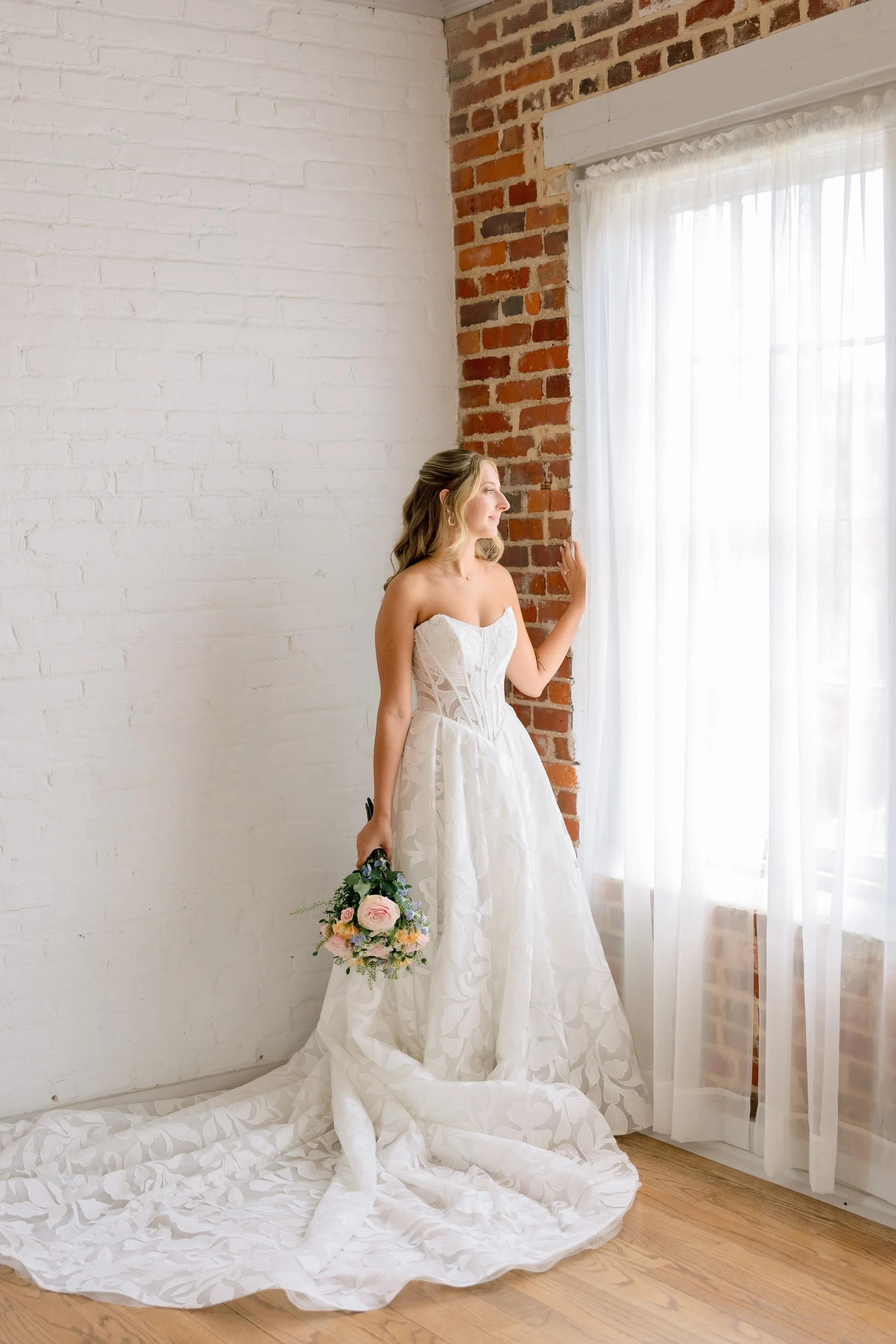 A woman in a wedding dress standing near a window with sheer curtains, holding a bouquet of flowers, with a long train of her dress spread out on the wooden floor.