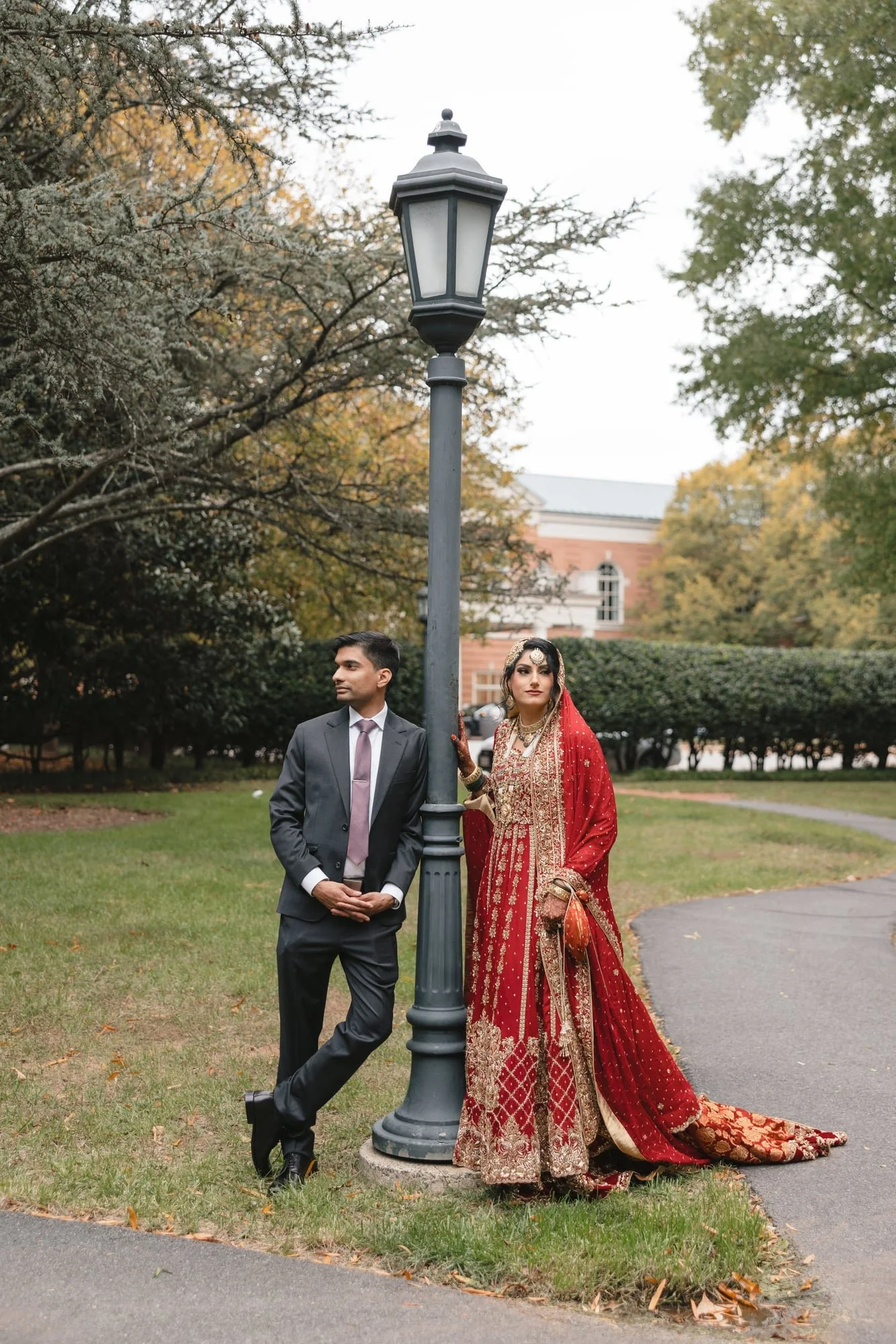 A man in a suit and woman in traditional Indian attire standing outdoors around a streetlamp in a park with trees and a building in the background.