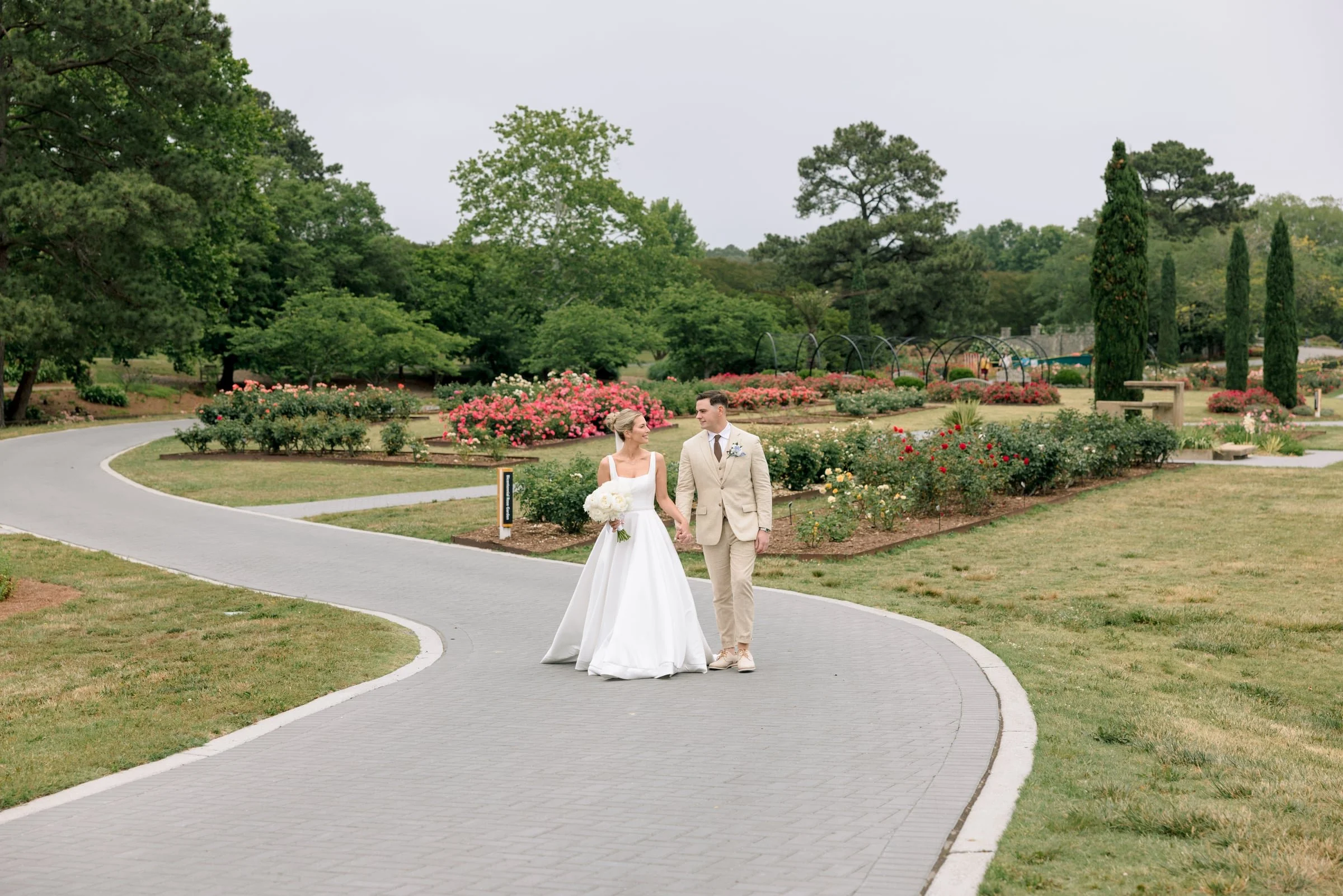 A bride and groom hold hands and walk along a paved garden path with blooming flowers and green trees in the background.
