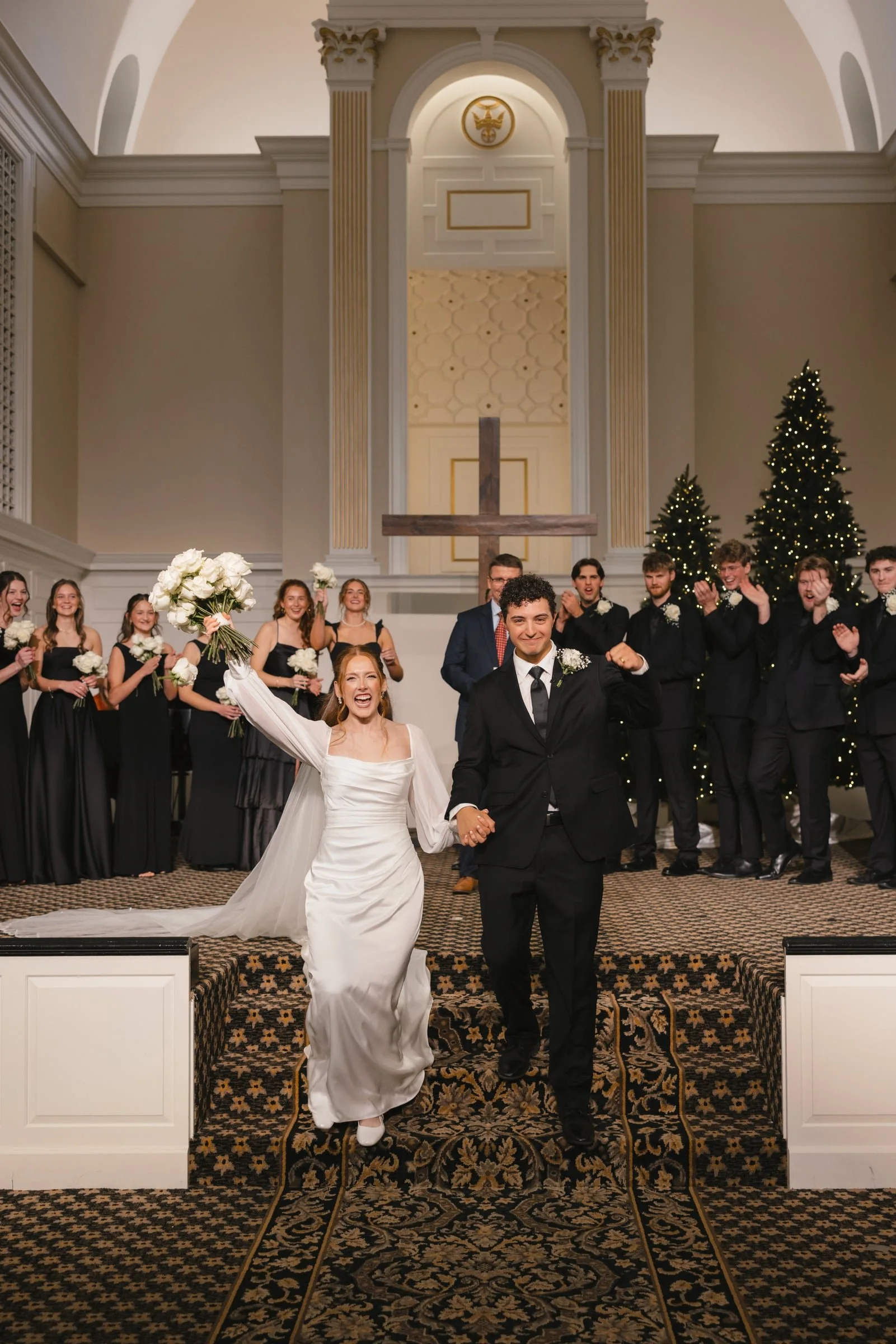 A wedding ceremony taking place in a church with the bride and groom standing in front of a cross, surrounded by bridesmaids and groomsmen, with Christmas trees and festive decorations in the background.