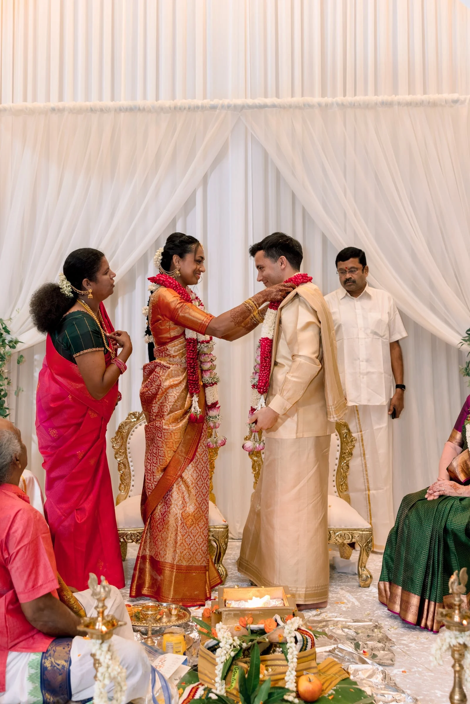 An Indian wedding ceremony with the bride and groom exchanging garlands, surrounded by family members. The bride is dressed in a traditional orange and gold saree, and the groom in a cream-colored traditional outfit. The backdrop features white curta
