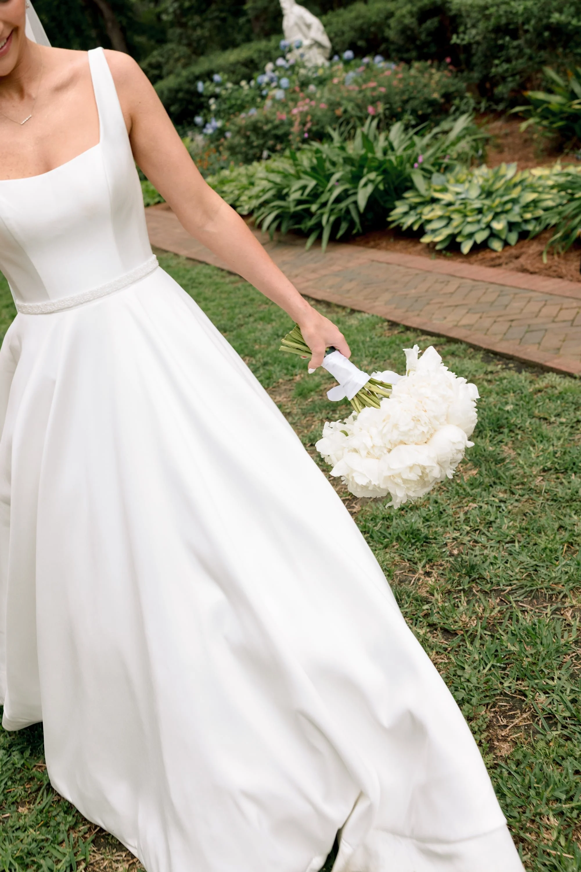 Close-up of a bride in a white wedding dress holding a bouquet of white flowers outdoors with green foliage and a brick pathway in the background.