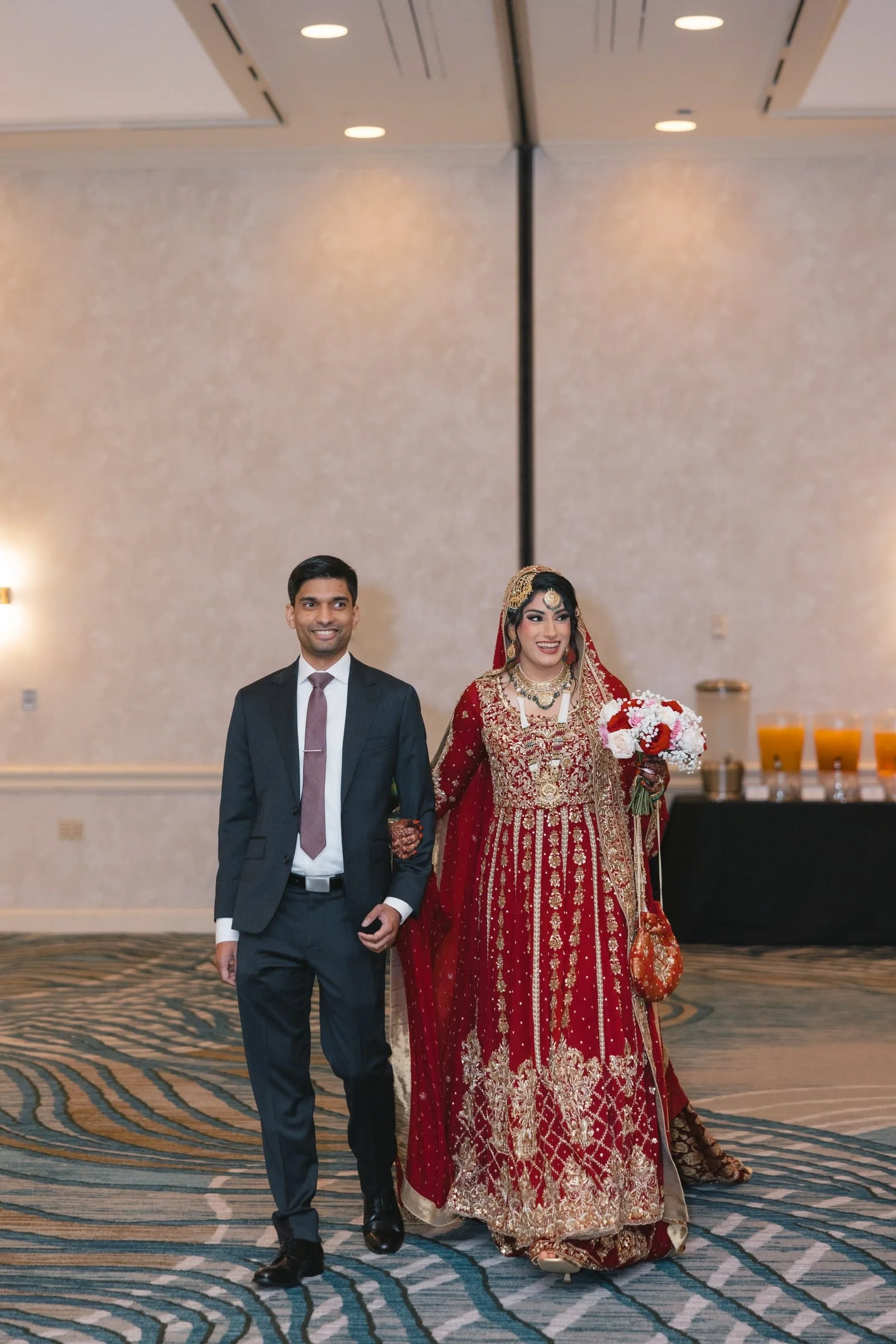 A couple walking hand in hand at a wedding reception. The bride is dressed in a traditional red and gold bridal outfit, holding a bouquet of flowers, and the groom in a dark suit with a tie. They are smiling and appear happy.
