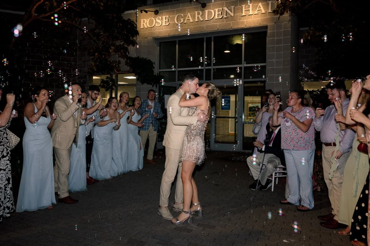 A bride and groom sharing a kiss in front of their wedding guests outside the Rose Garden Hall, with guests clapping and taking photos, and decorative lighting and bubbles in the scene.