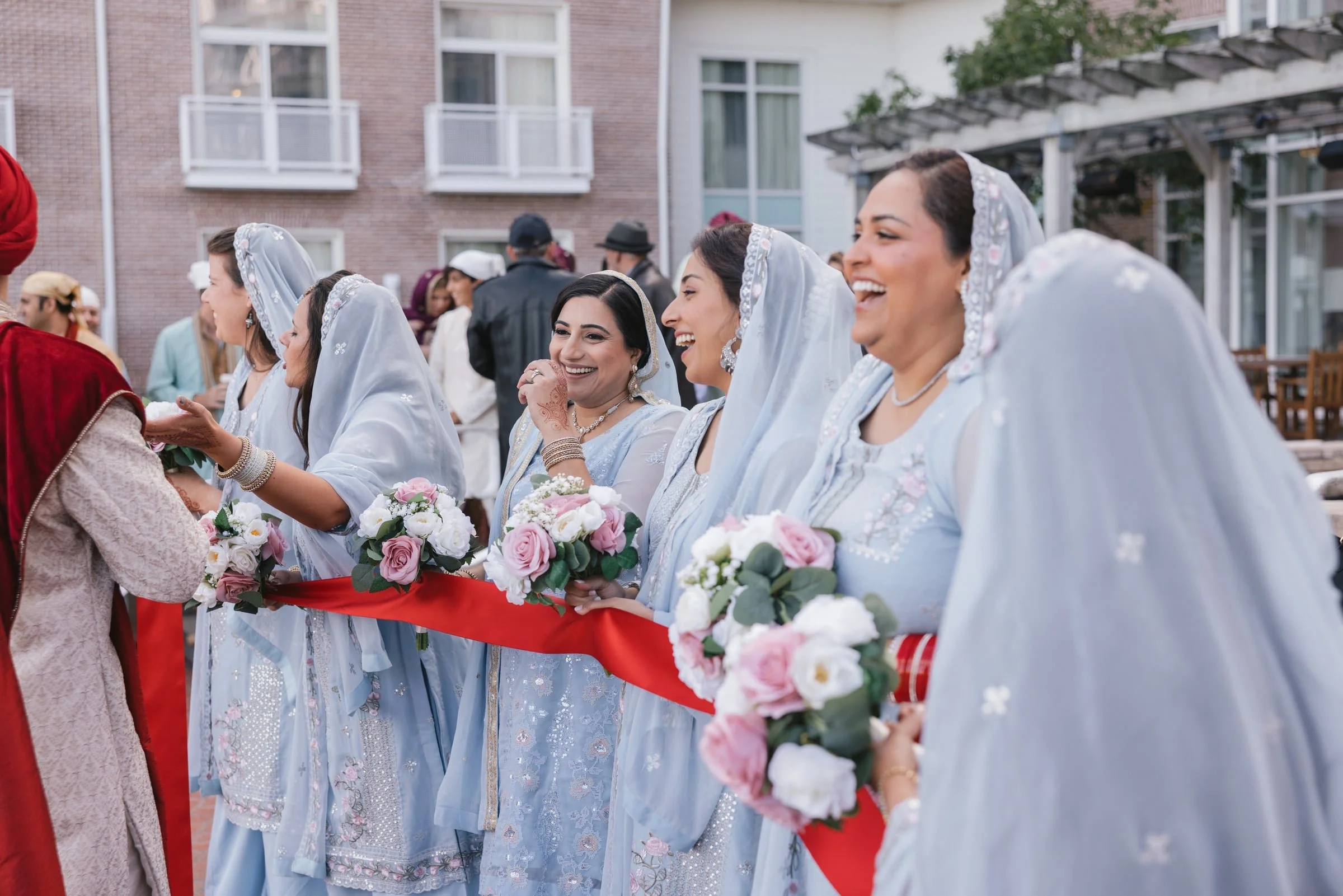 A group of women dressed in traditional South Asian attire, holding bouquets of pink and white roses, participating in a wedding ceremony outdoor in front of a modern building.