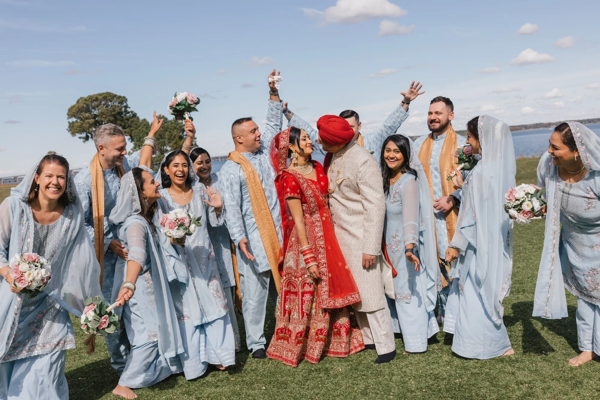 A group of people dressed in traditional South Asian attire, celebrating outdoors by a body of water on a sunny day. The group includes men and women, some women wearing white and silver dresses with veils, and some holding bouquets. The couple in th