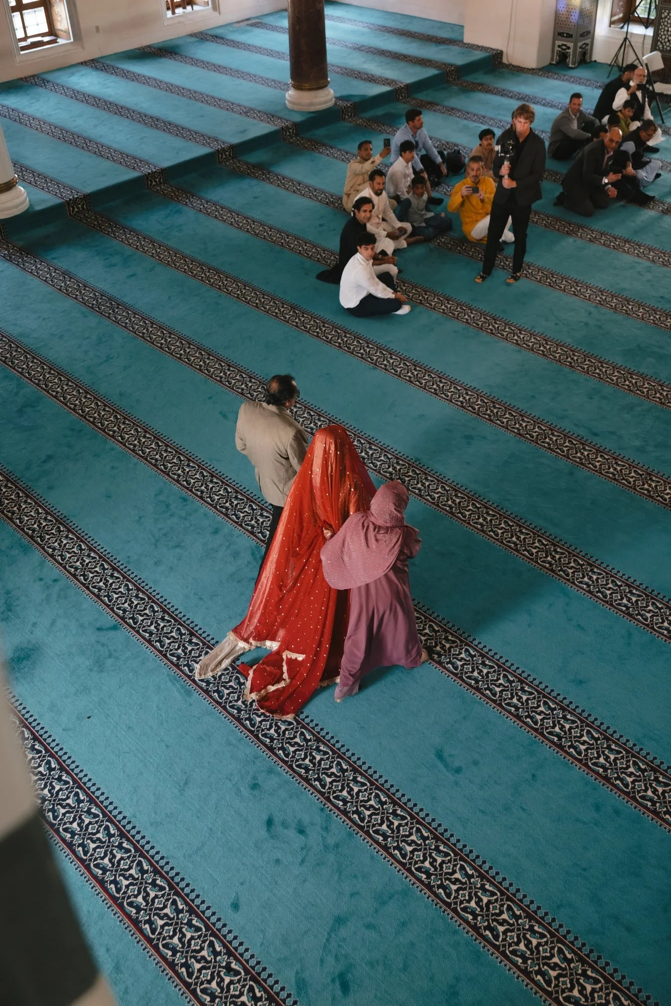 Two women in traditional Indian attire walking on a prayer rug inside a mosque, with men sitting on the rug in prayer or listening to a sermon nearby.