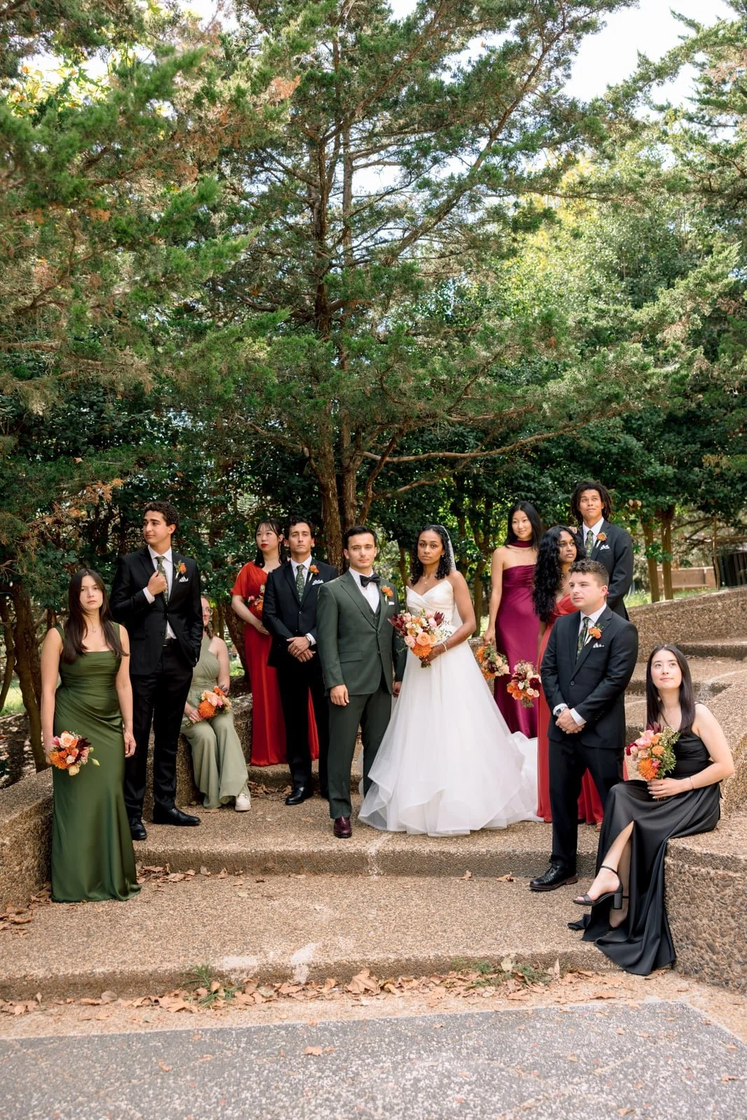 Group of people dressed in formal attire, including a bride in a white wedding gown and a groom in a green suit, standing outdoors on steps with trees in the background, holding bouquets of flowers.