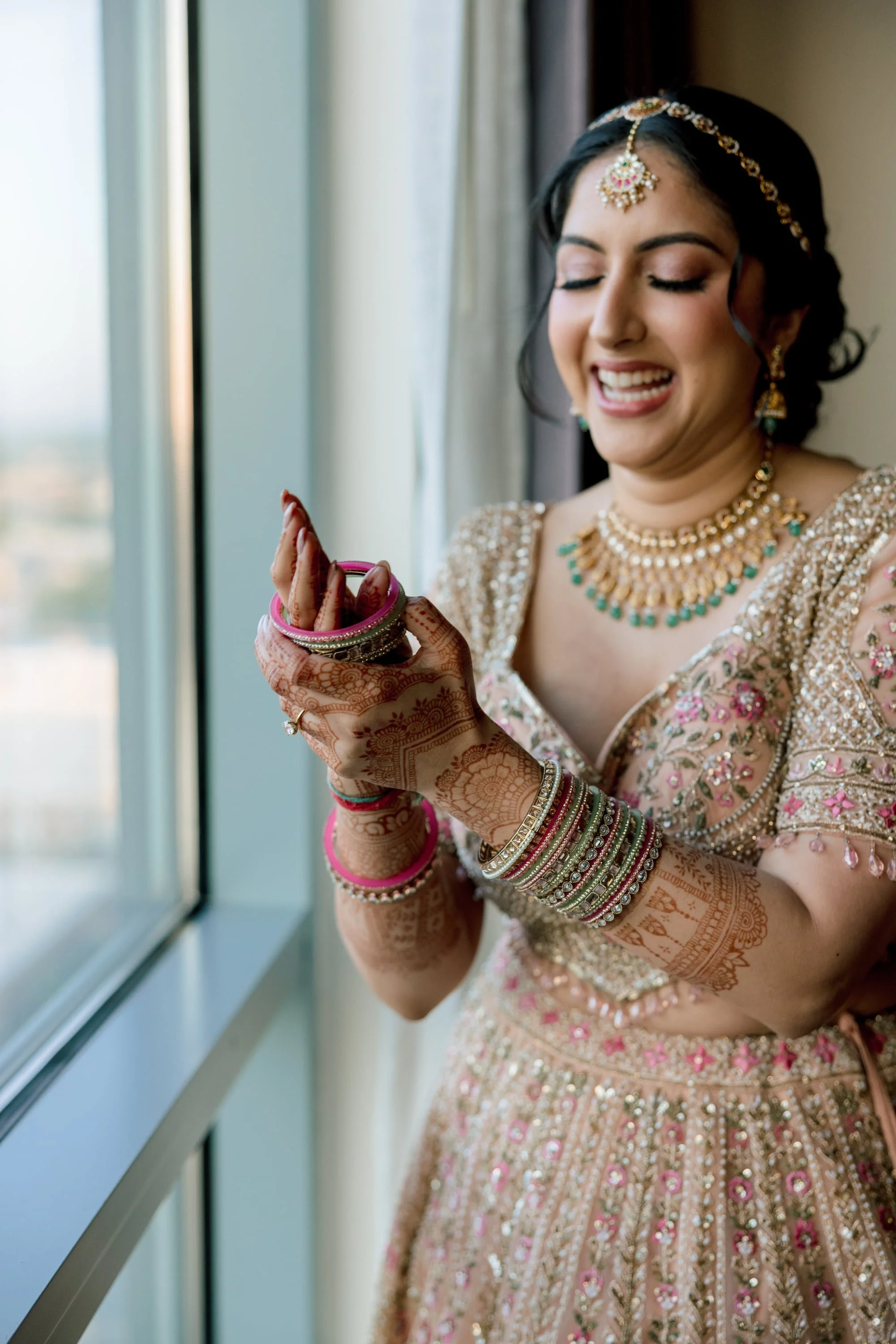 A woman dressed in traditional Indian bridal attire, wearing jewelry and henna on her hands, looking at her reflection near a window.