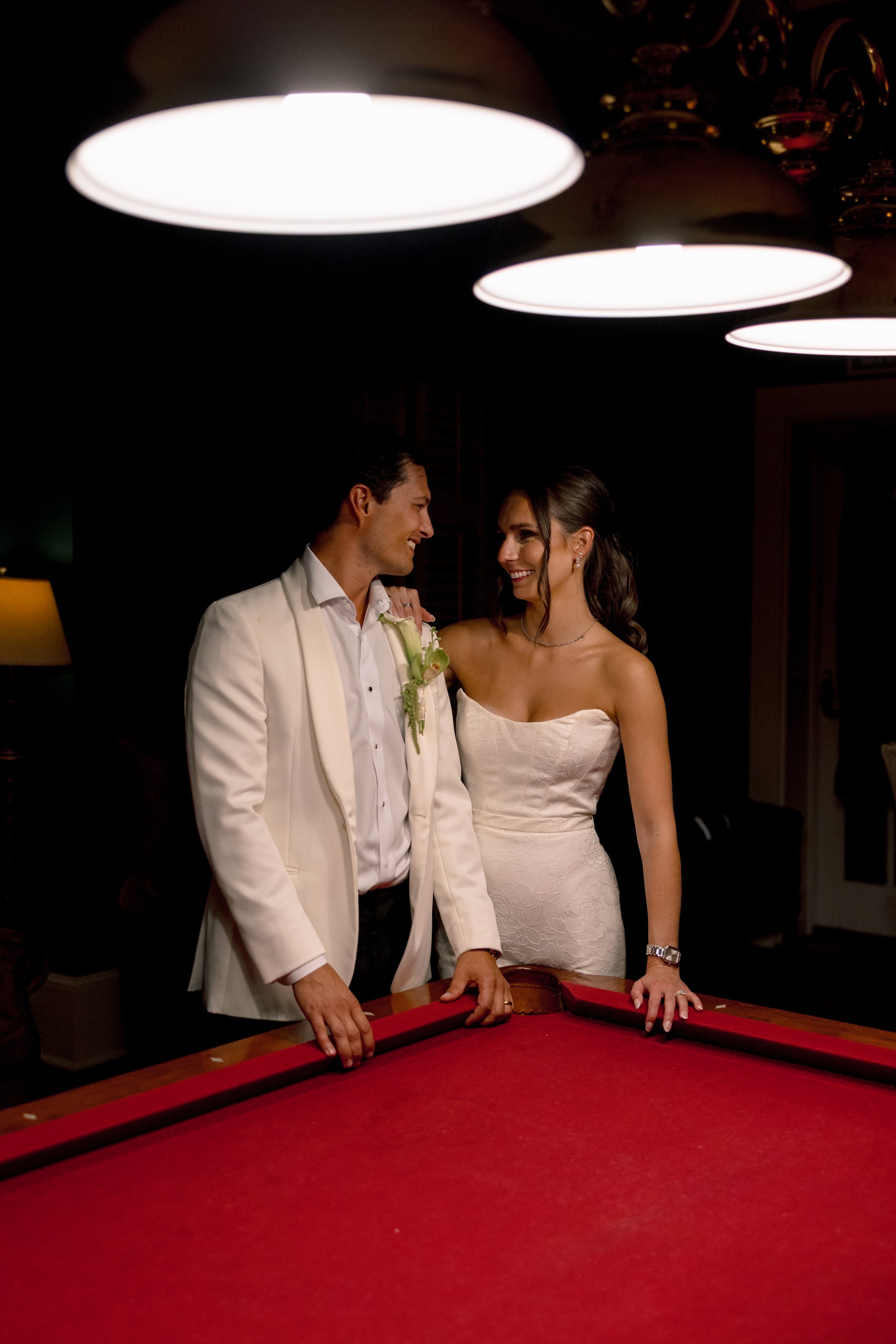 A man and woman dressed in formal attire standing together around a red felt pool table, smiling at each other in a dimly lit room with large ceiling lights overhead.