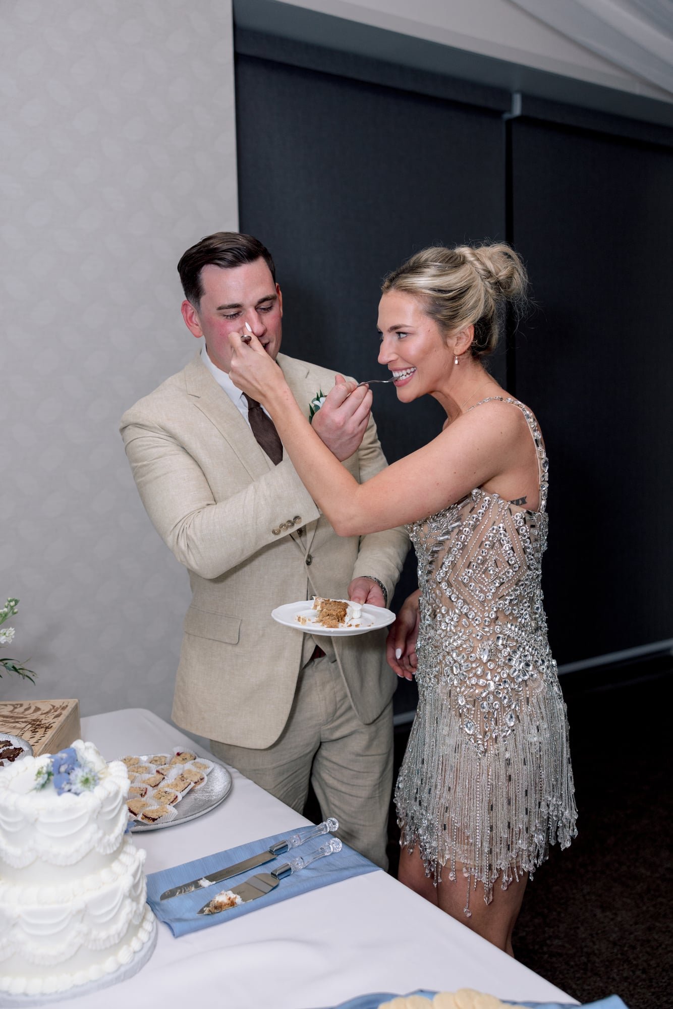 A couple at a wedding reception being served a piece of wedding cake. The bride is in a detailed, shiny dress and the groom in a beige suit. A table with a large white wedding cake decorated with flowers and plates of snacks is in front of them.