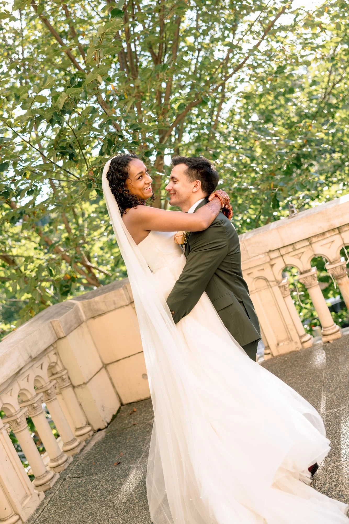 A bride and groom smiling and embracing outdoors, with greenery and stone railing in the background.