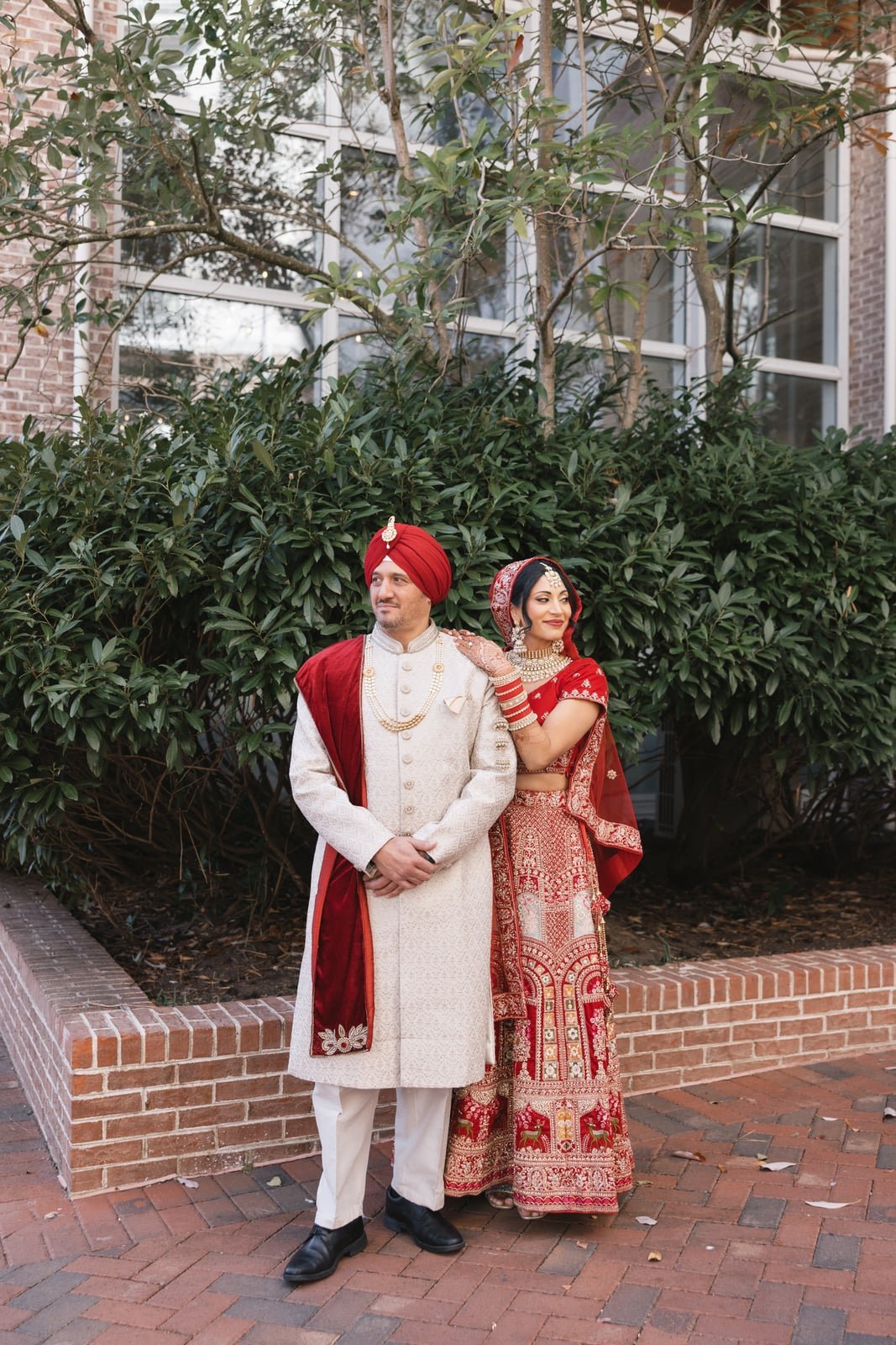 A bride and groom in traditional Indian wedding attire standing outdoors in front of lush green bushes and a building with large windows.