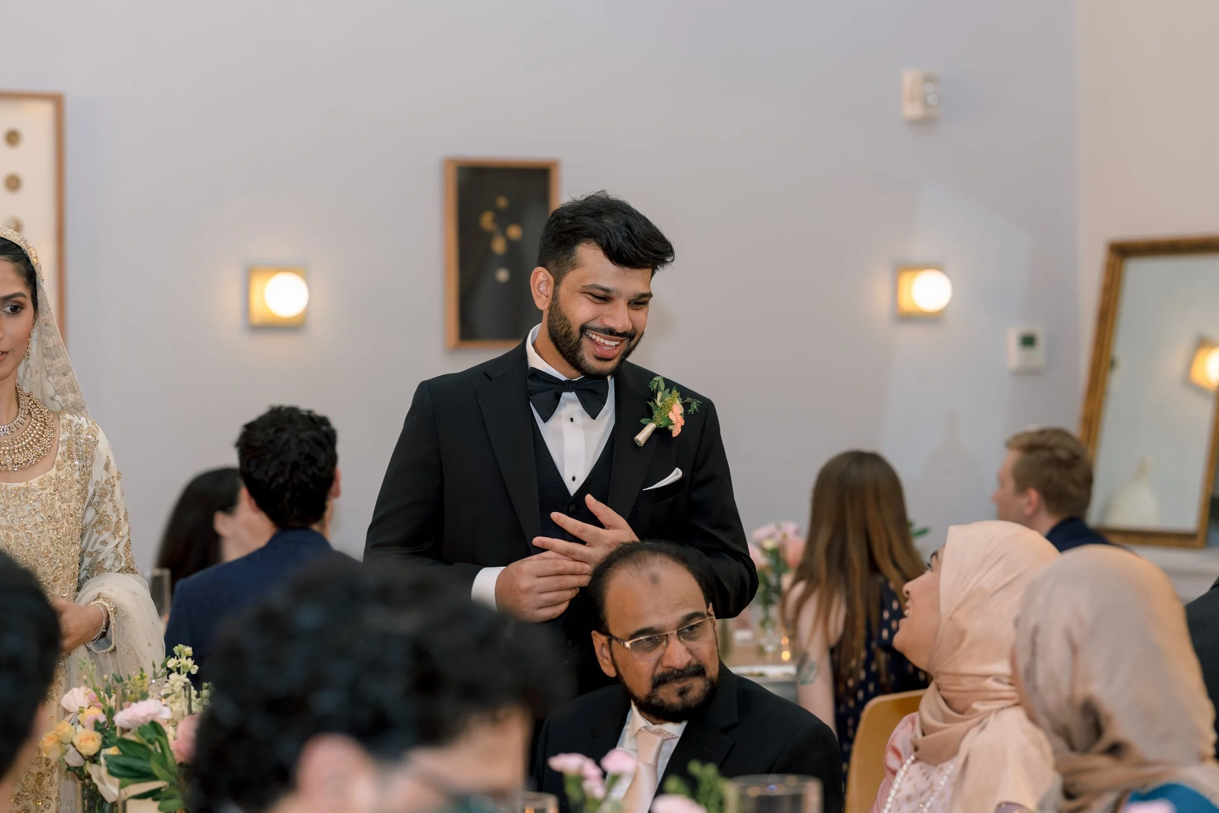 A groom in a black tuxedo with a bow tie and flower boutonniere smiling and talking to guests at a wedding reception. Female guests in traditional South Asian attire are also visible, seated at decorated tables with flowers.